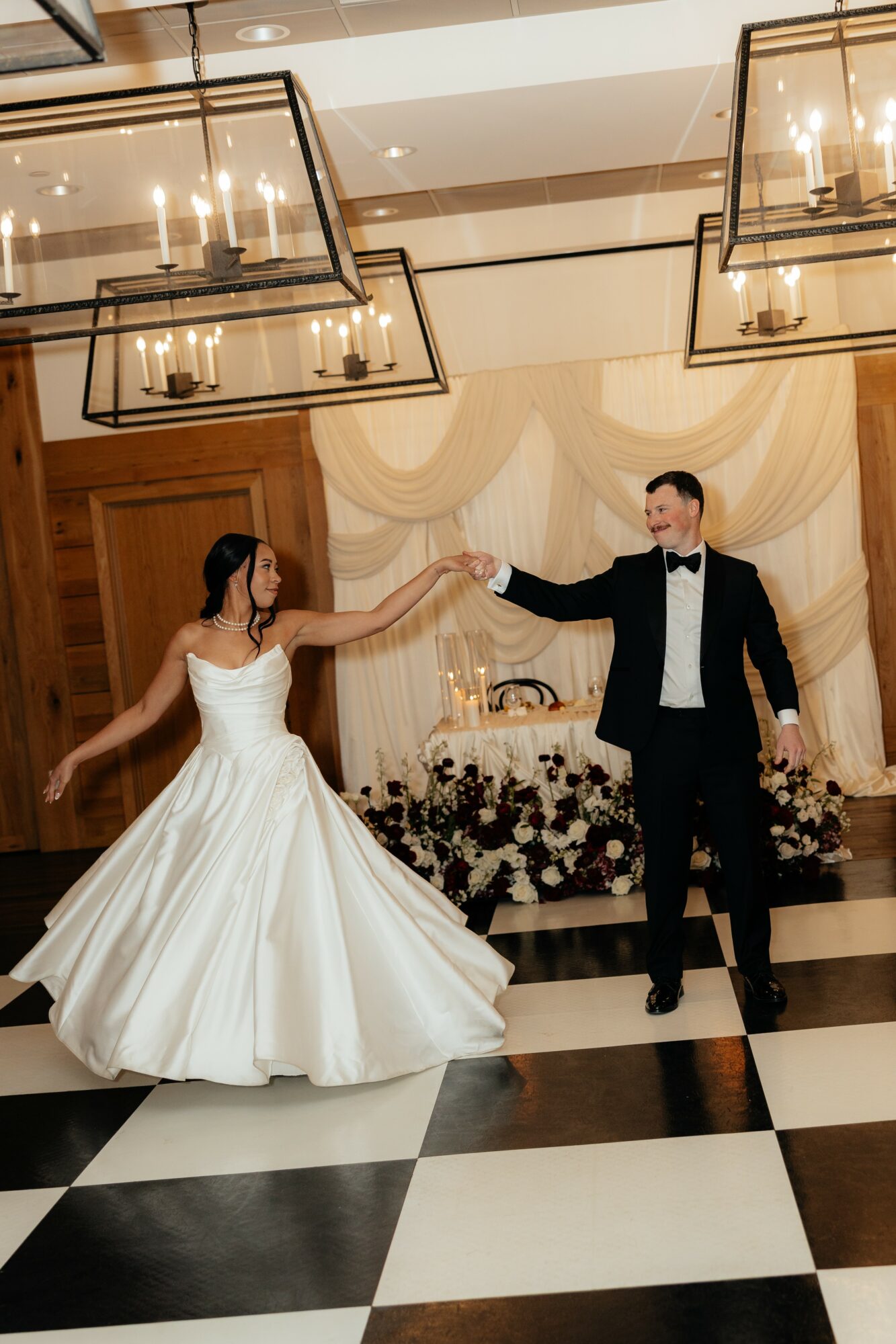 Bride and groom dancing indoors, holding hands, with wedding decorations and a table in the background.