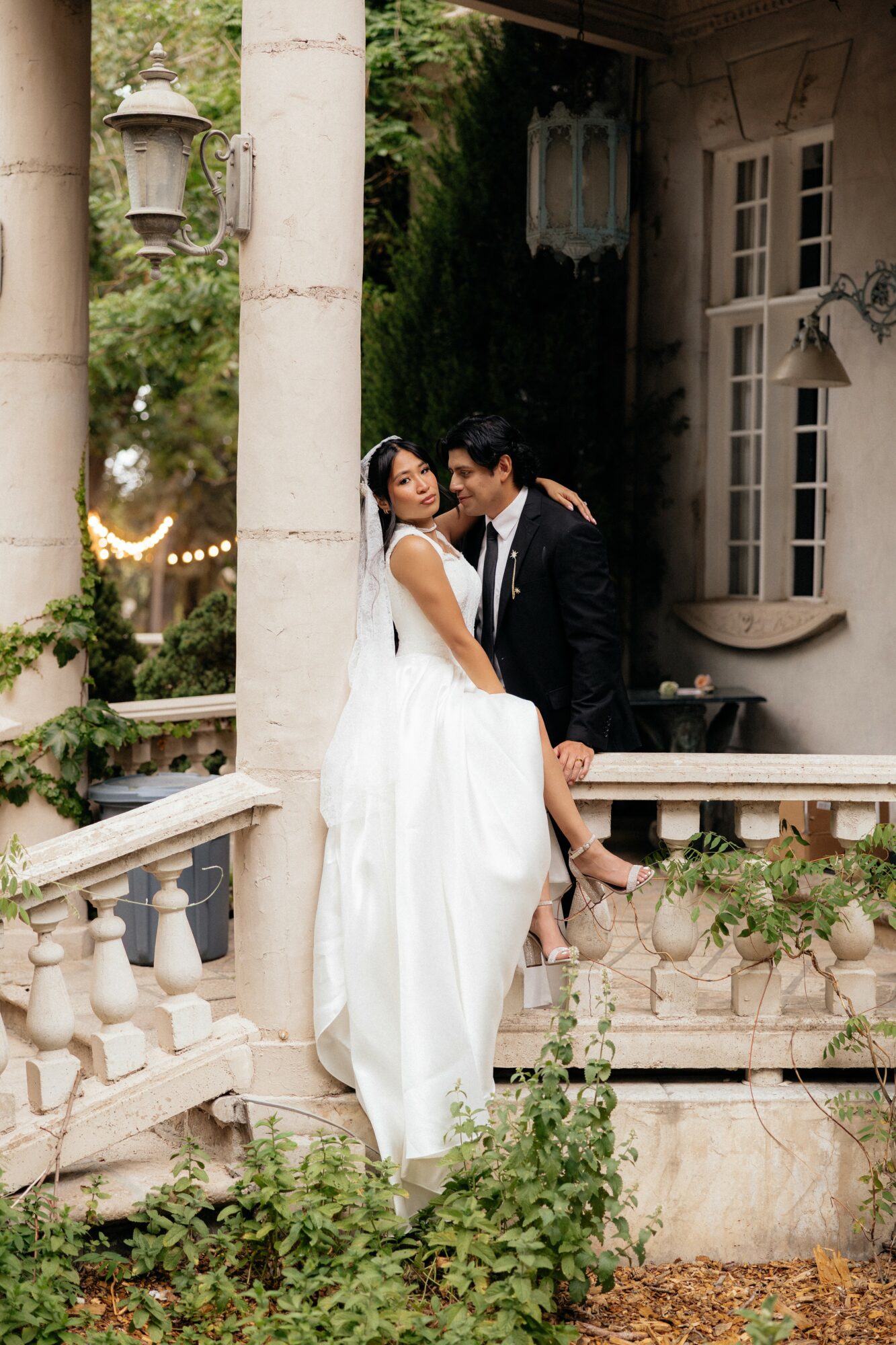 A bride and groom pose on a balcony with greenery and a building in the background.