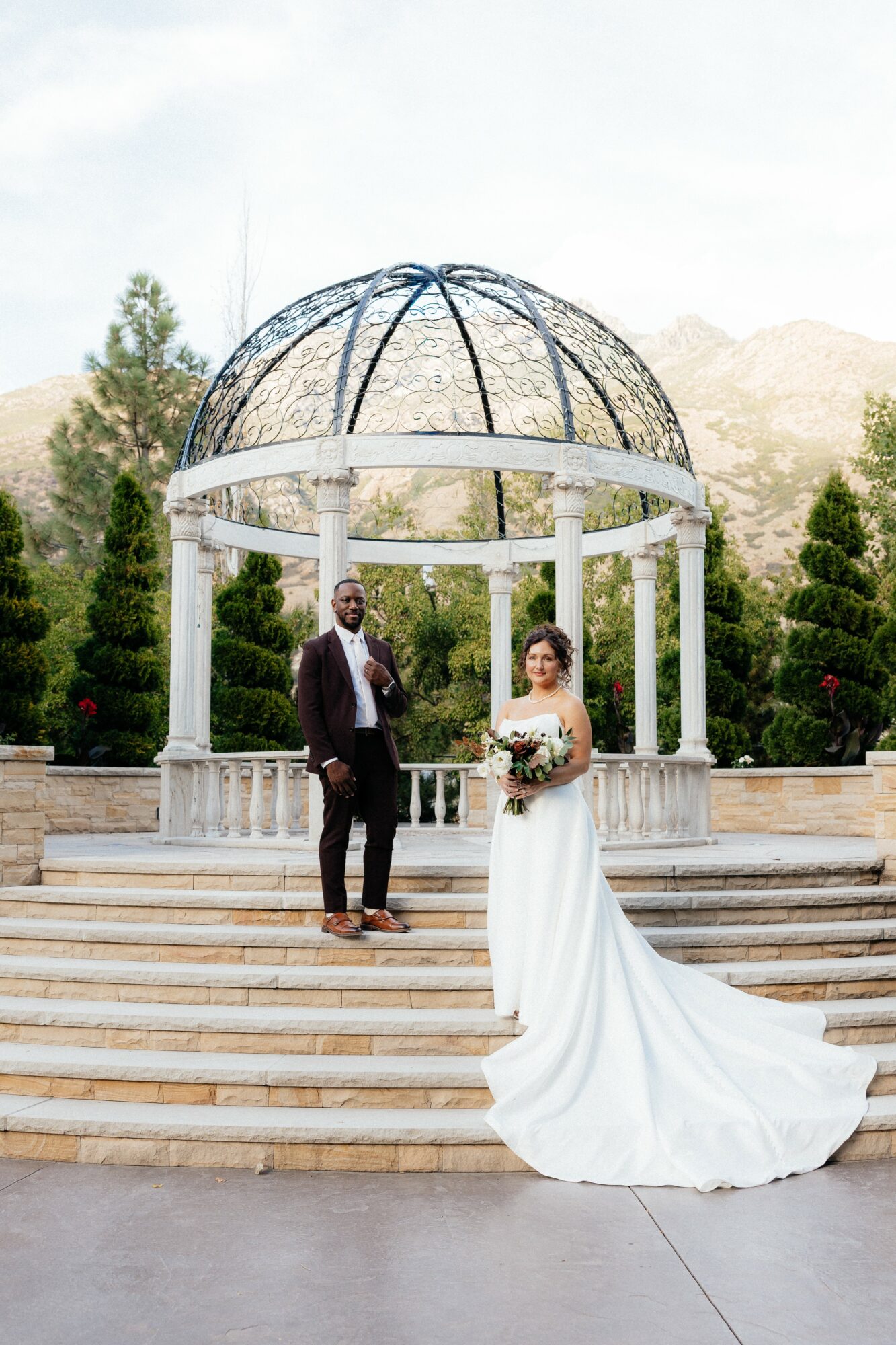 Bride and groom stand on steps under a decorative gazebo outdoors, with trees and mountains in background.
