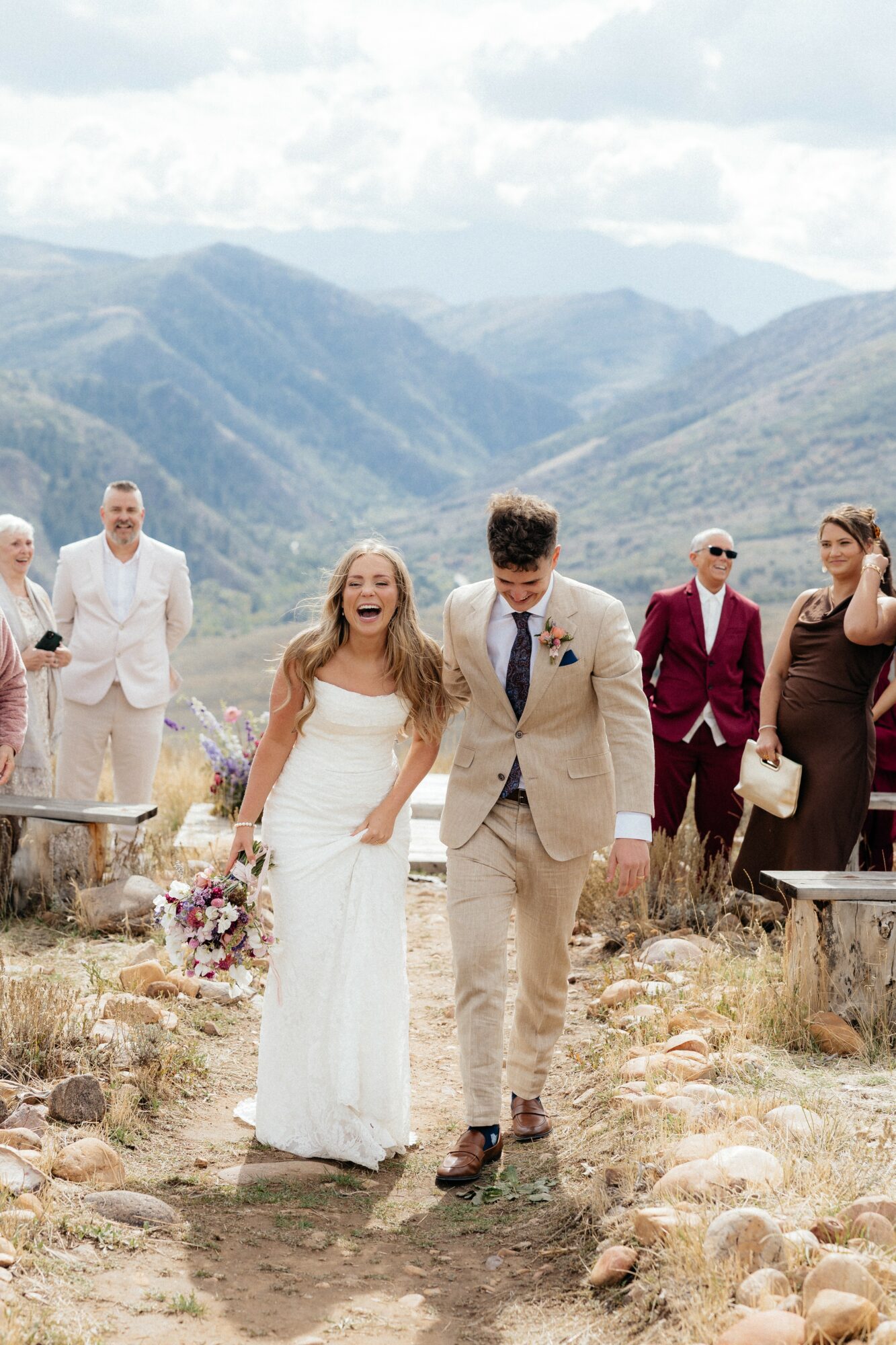 Bride and groom walking outdoors with mountains in background, smiling, wedding guests behind them, rocky ground, cloudy sky.