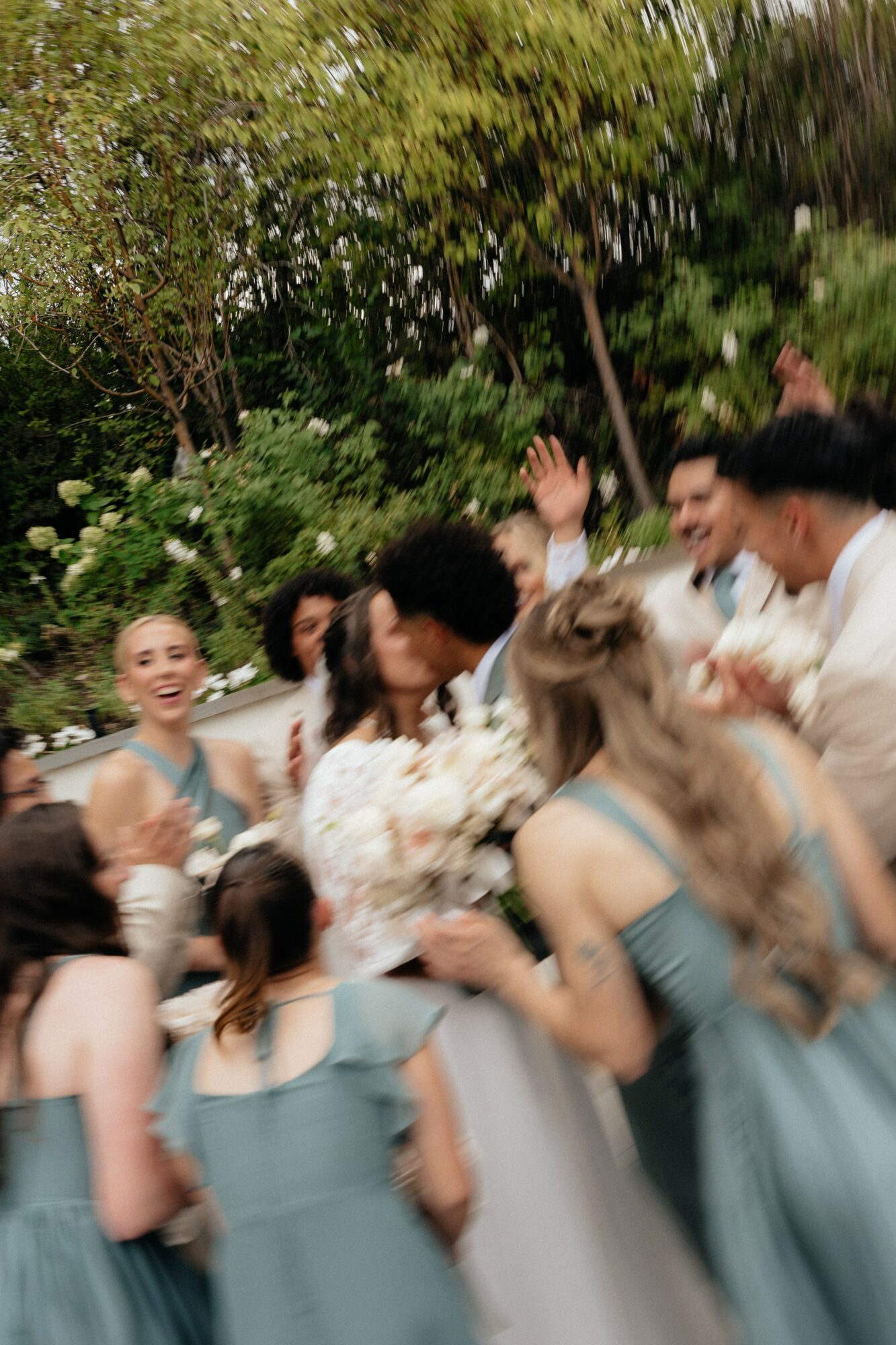 Group of people celebrating outdoors, smiling, raising hands, surrounded by greenery, wearing light-colored and blue clothing.