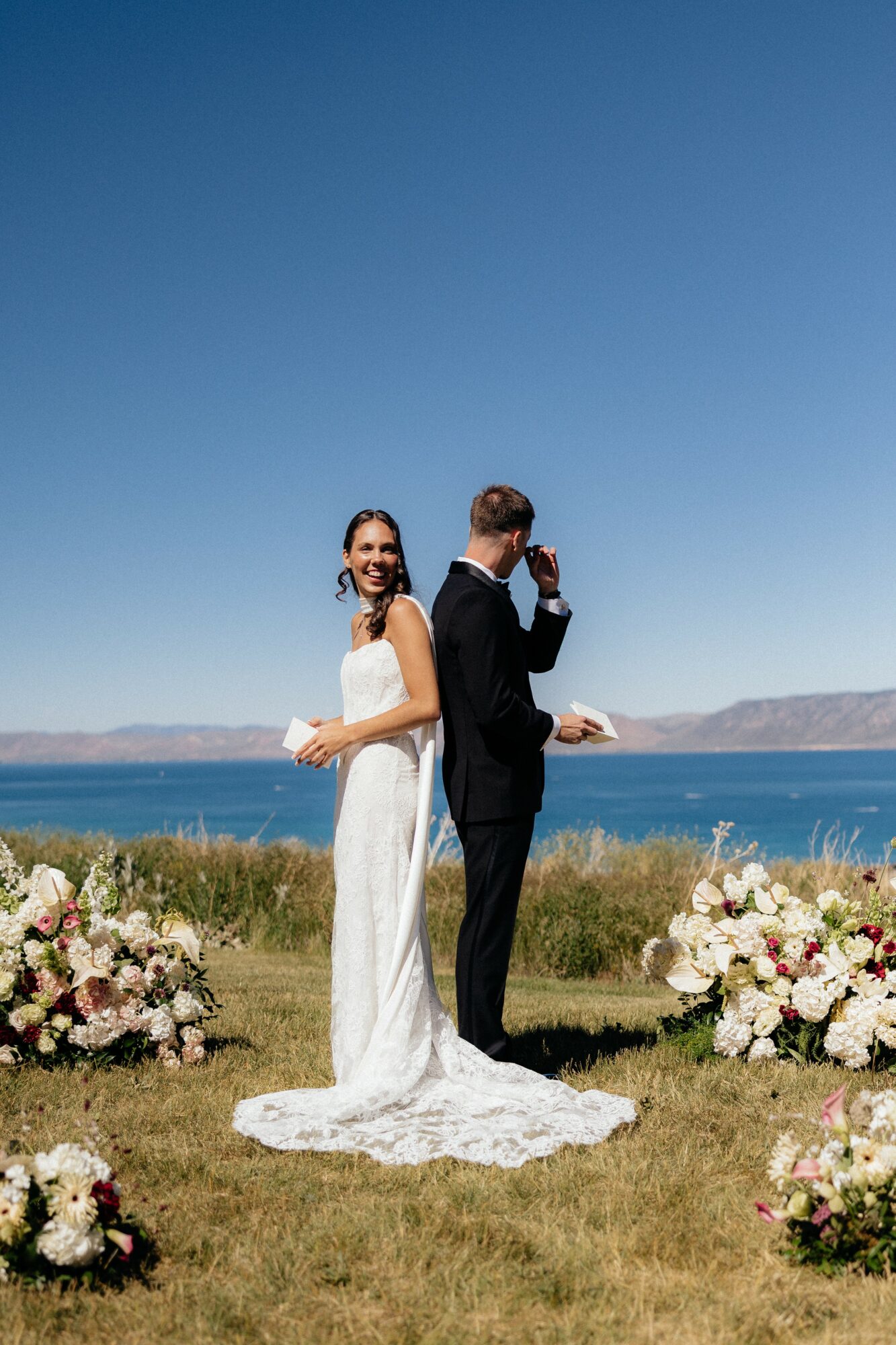 A bride and groom stand outdoors near water with flowers around them, under a clear blue sky.