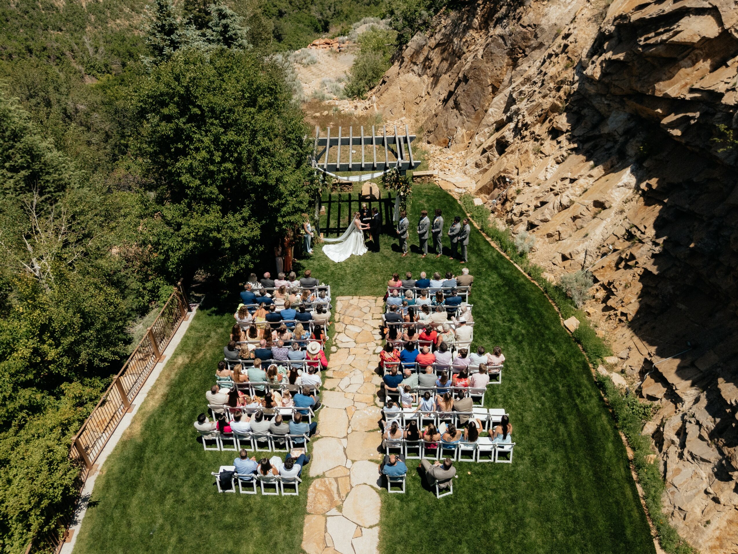 Outdoor gathering with chairs arranged in rows on grass, surrounded by trees and rocky cliffs, with a pathway in the center.
