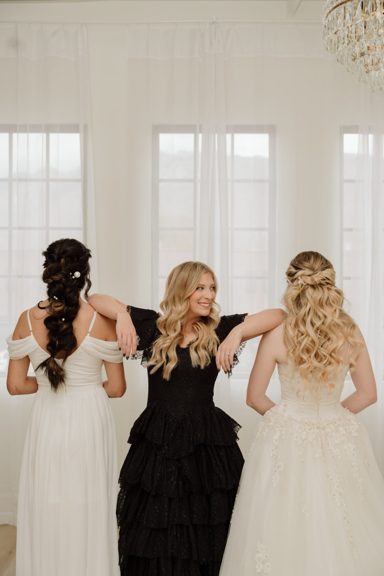Three women in wedding dresses standing in a bright room with large windows, facing away from the camera, with one woman in the middle smiling.