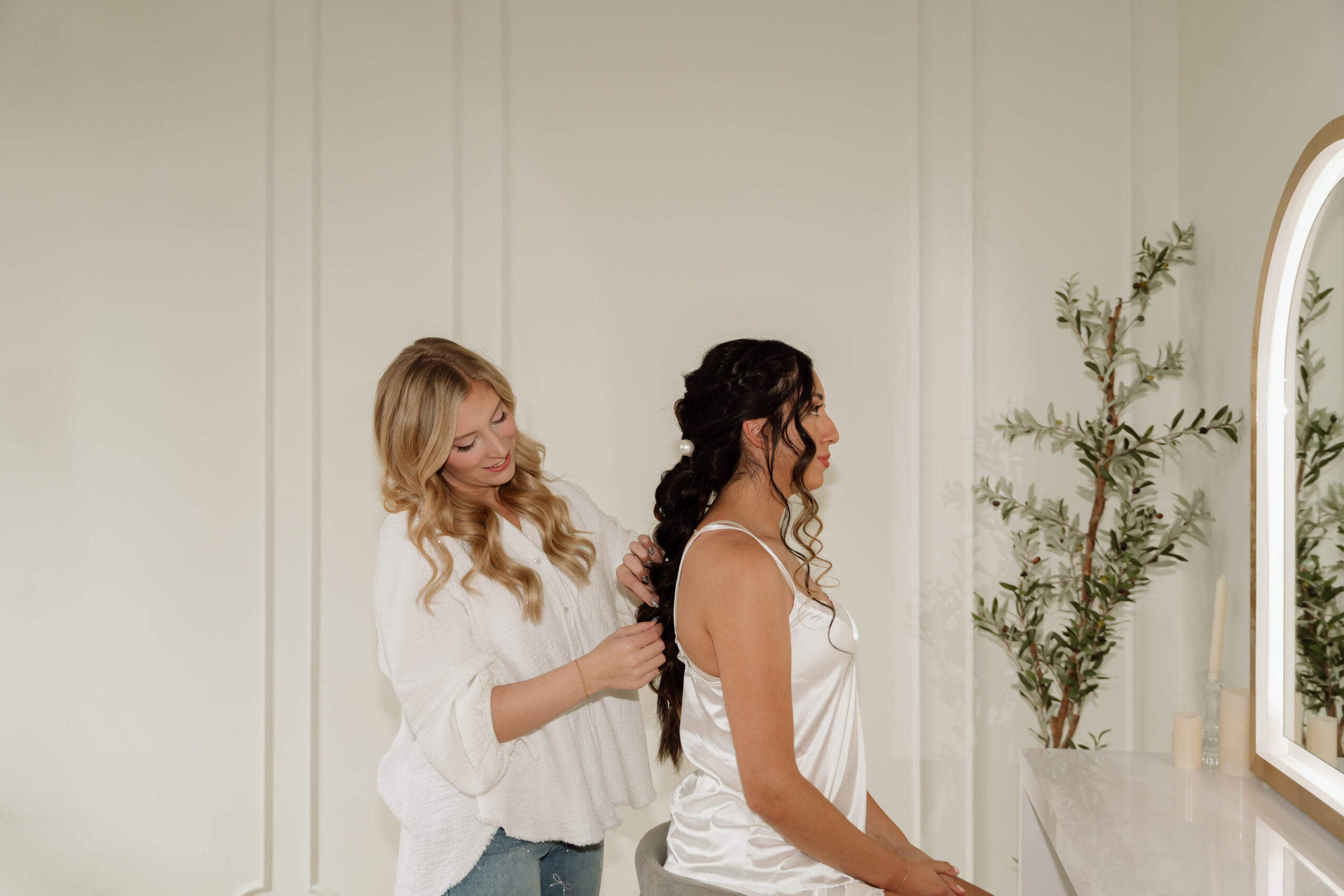 Two women with long hair, one with blonde and one with dark hair, in a bright room with a mirror and plant.