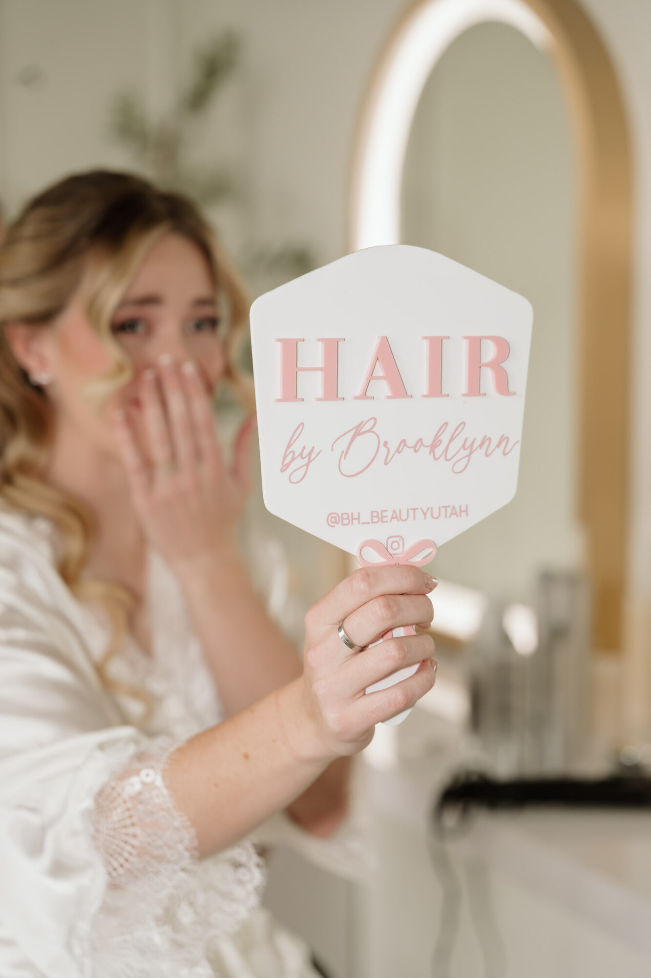 Woman holding a mirror with pink text reading 'HAIR by Brallyn' in a salon, blurred background.