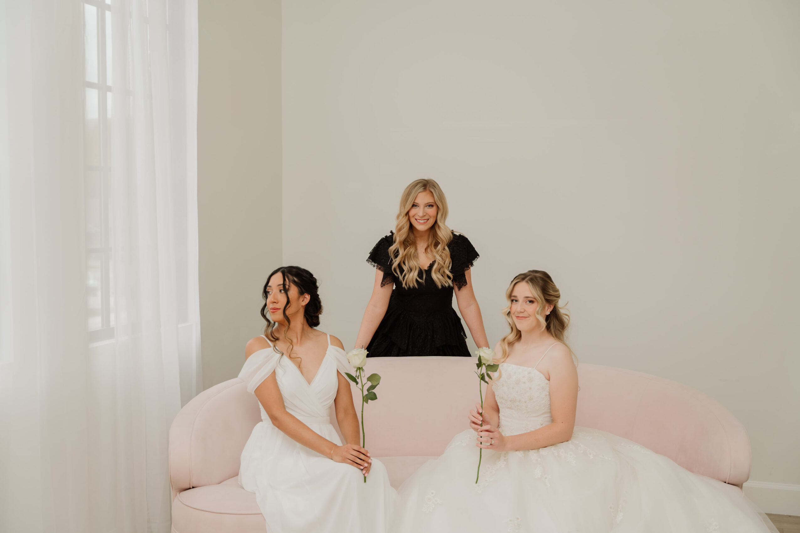 Three women in white dresses, two sitting on a pink sofa, one standing behind, in a bright room with large window.