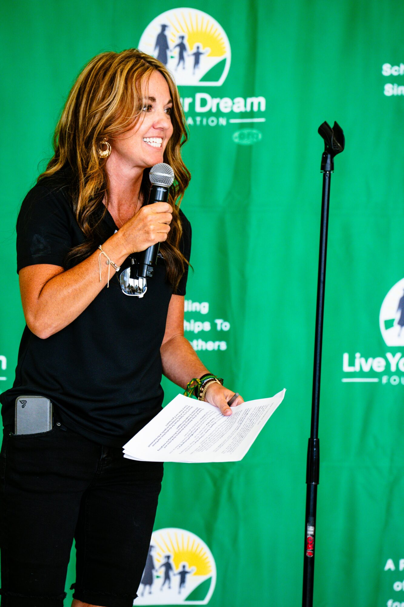 Woman with long hair holding a microphone and paper, standing in front of a green backdrop with logos.