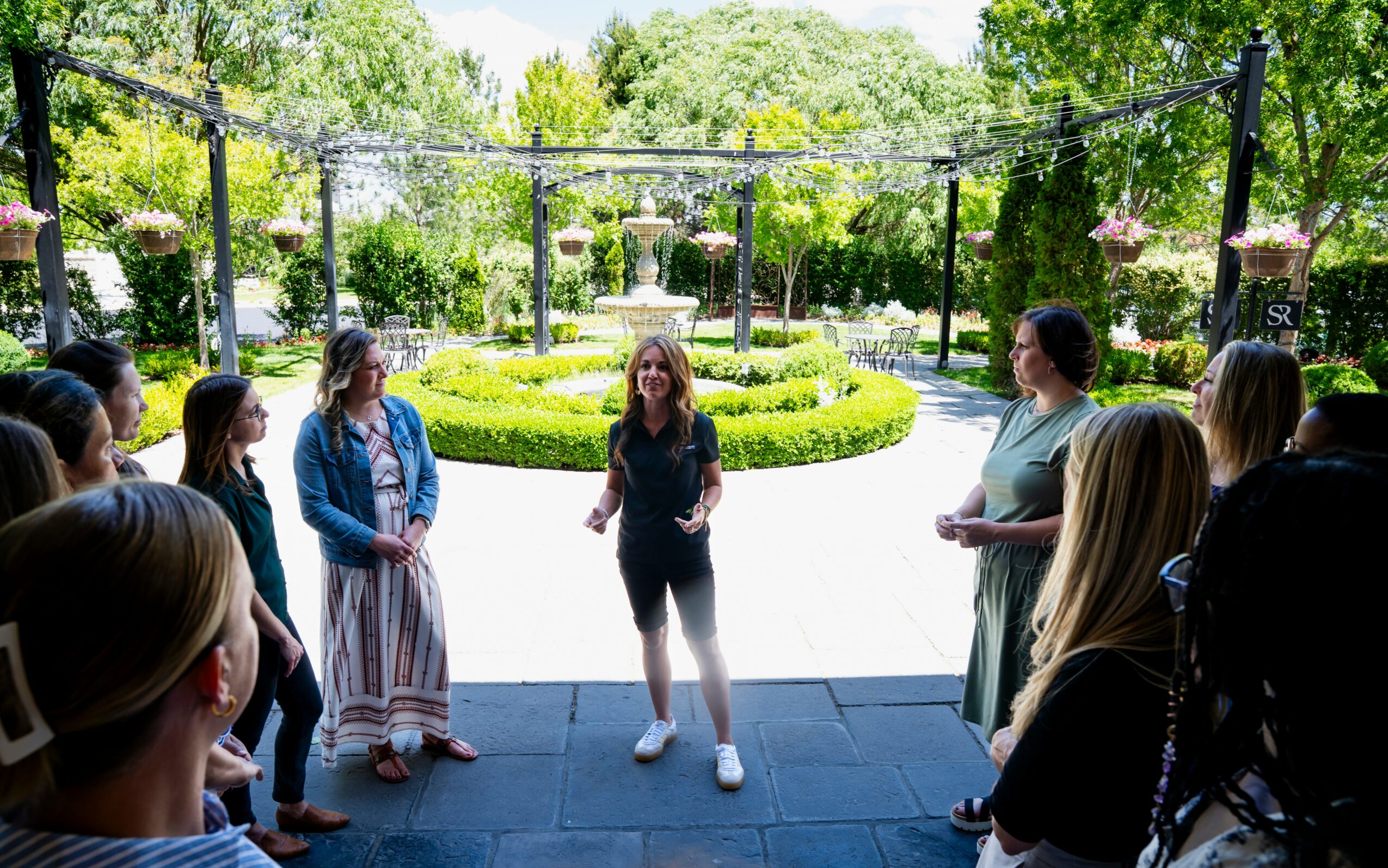 Group of people gathered outdoors in a circle around a woman speaking, with a garden and trees in the background.