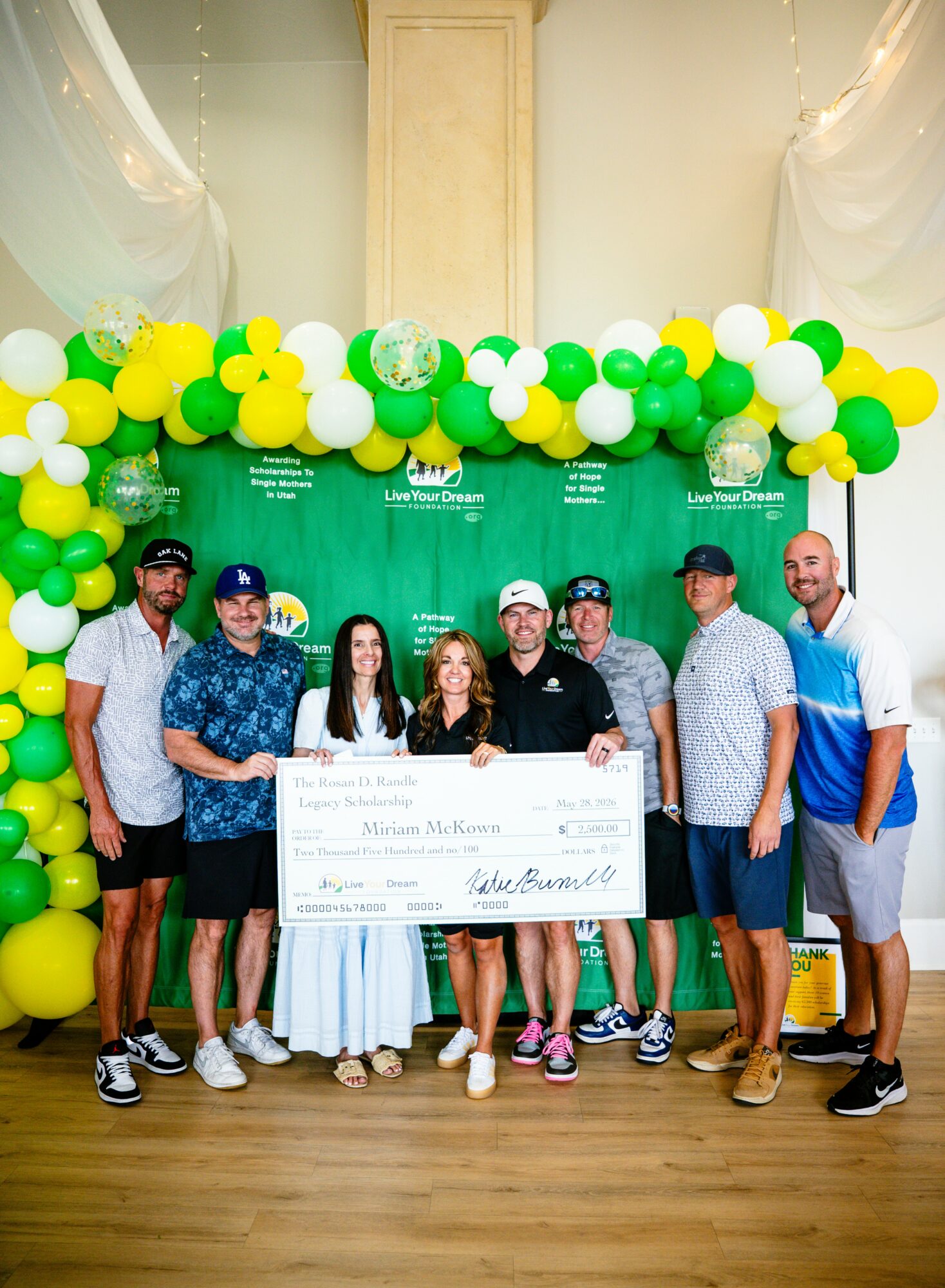 Group of nine people holding a large check, standing in front of a green backdrop with yellow, white, and green balloons, indoors.