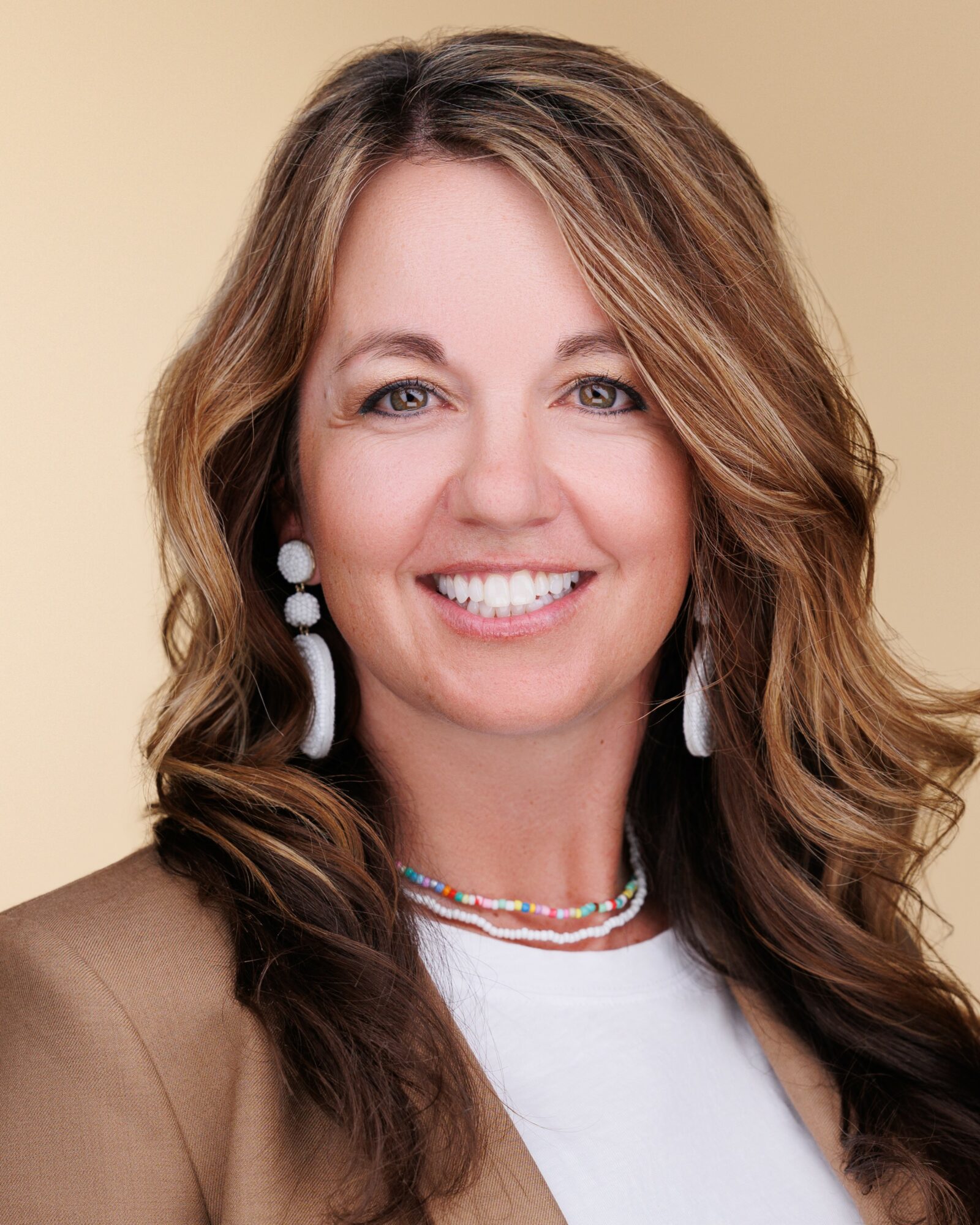 Woman with long wavy hair smiling, wearing earrings and a necklace, against a light background.
