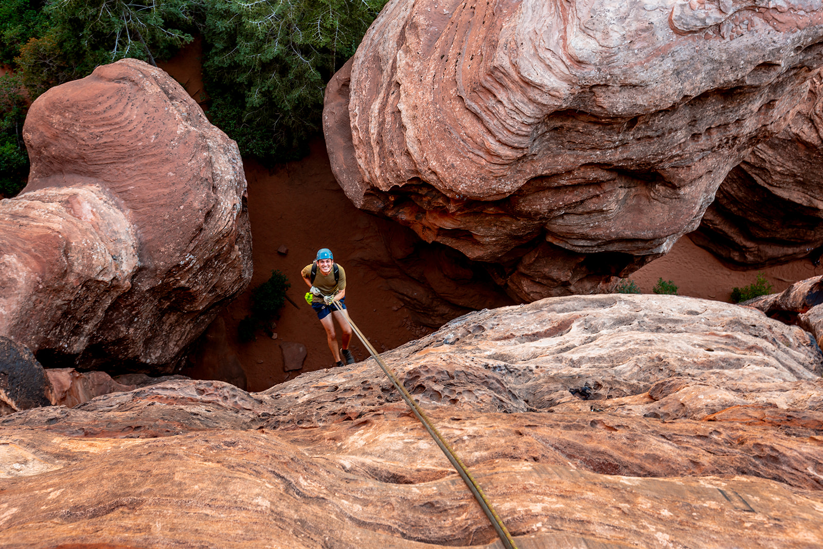 Person wearing helmet and harness rappelling down a rocky cliff with large boulders around.