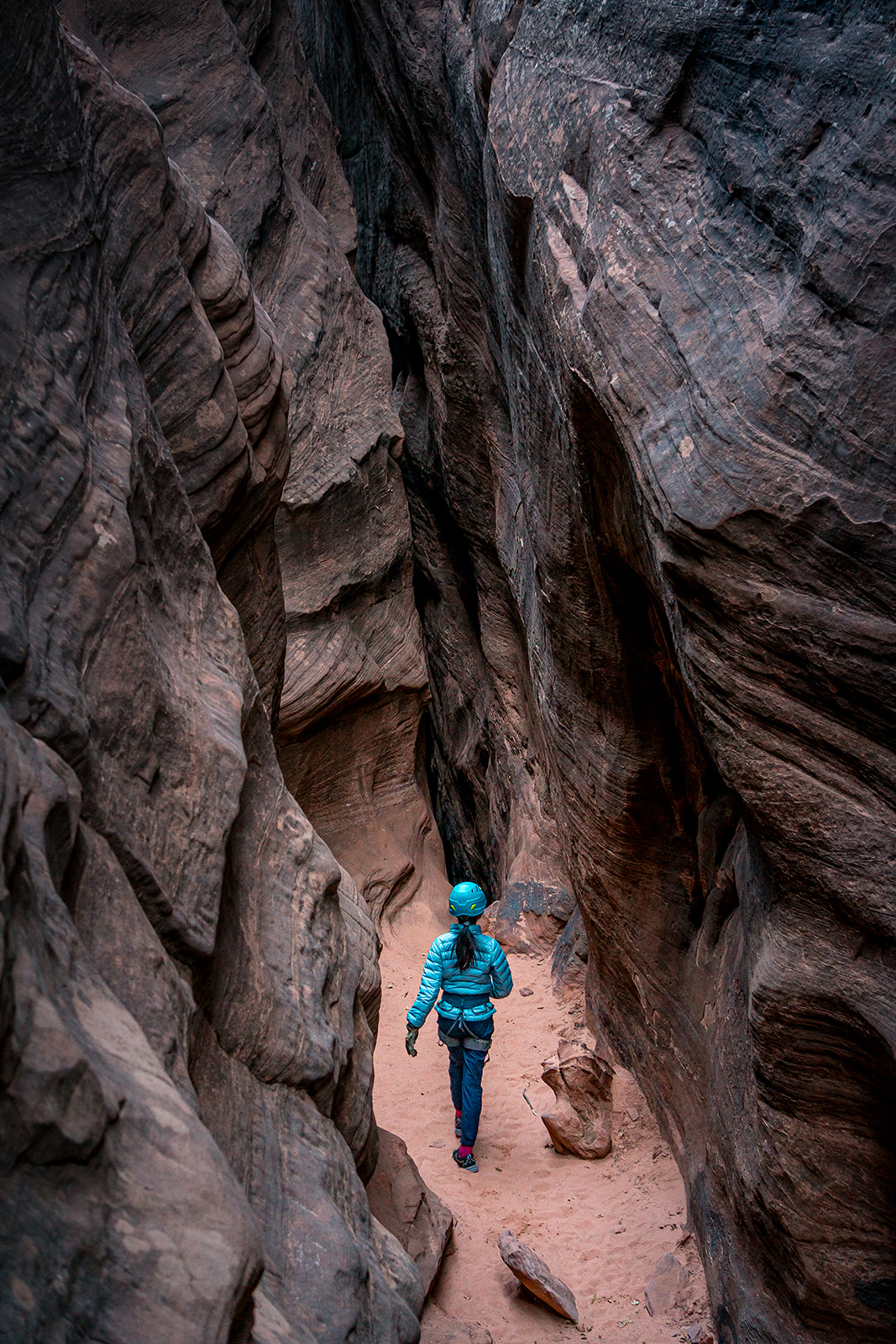 Person wearing a blue jacket and helmet walking through a narrow canyon with tall rock walls.