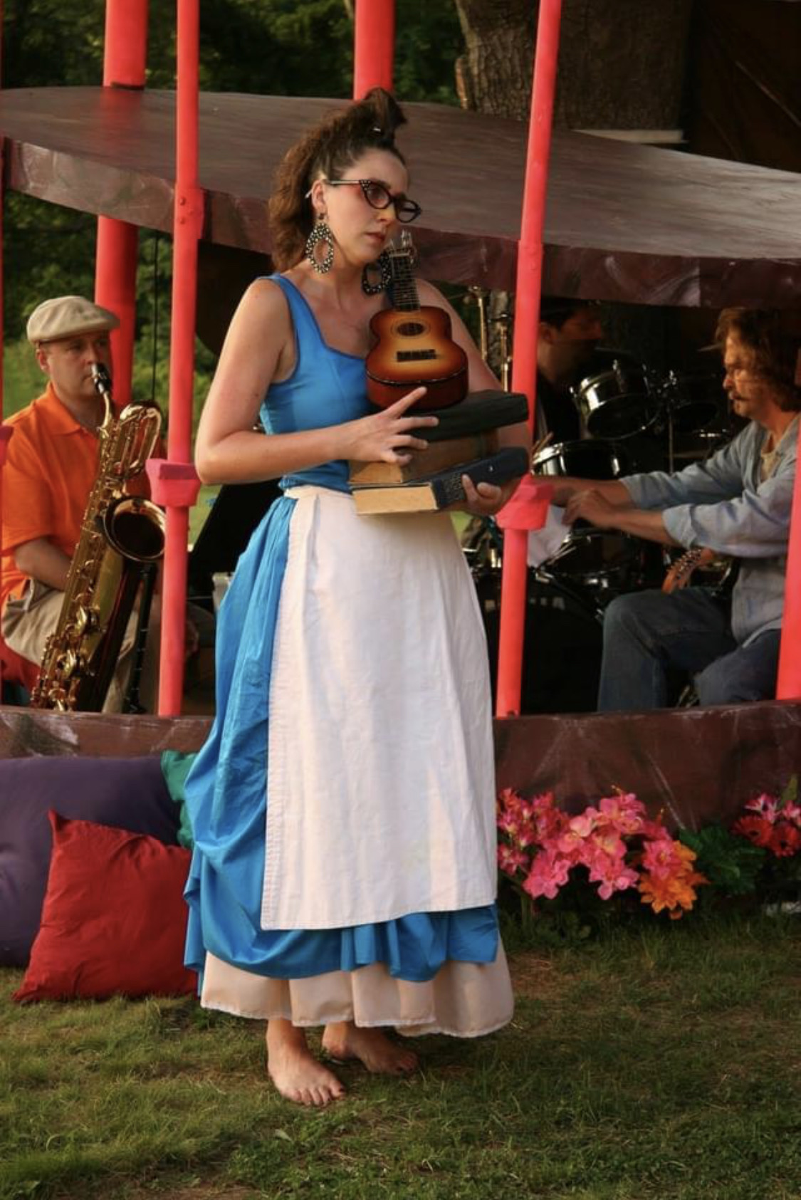 Woman in blue dress holding a guitar, standing outdoors with musicians and flowers around her.