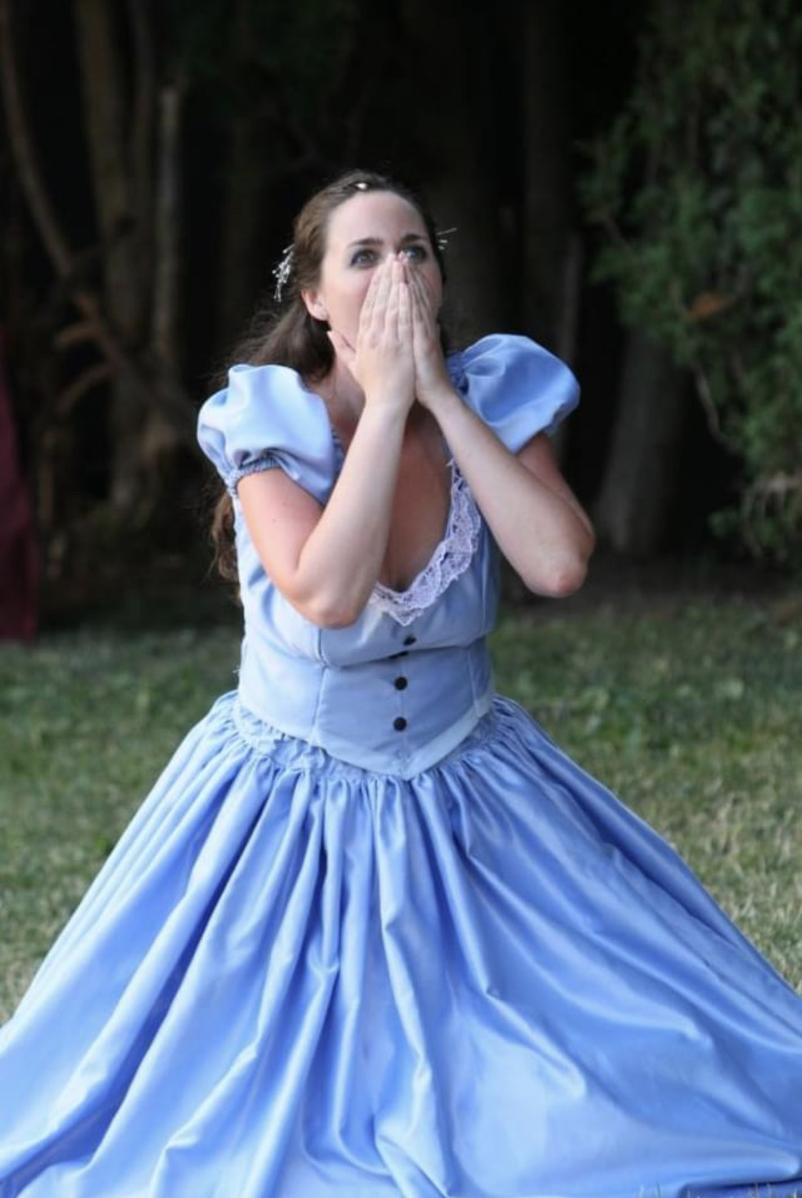 Woman in a blue dress covering her mouth with both hands outdoors, surrounded by trees and grass.