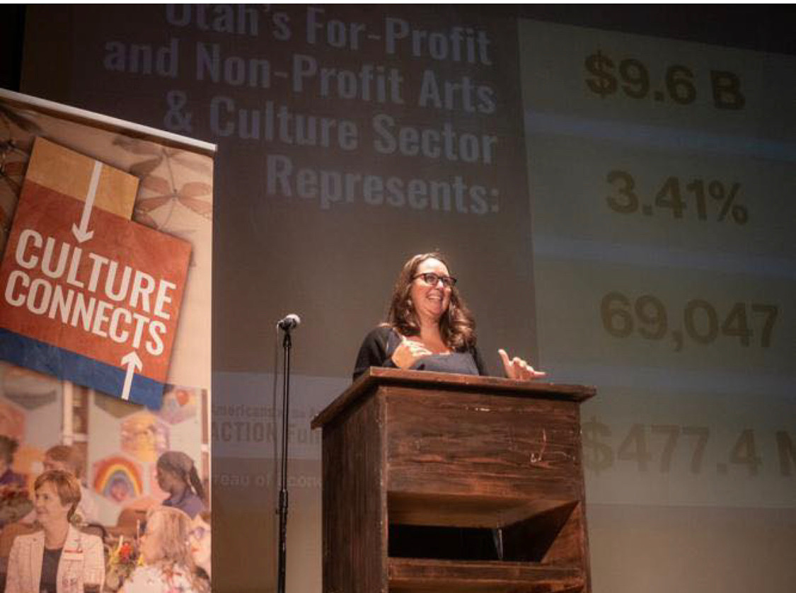 Woman speaking at a podium during a presentation with a slide showing financial data in the background.