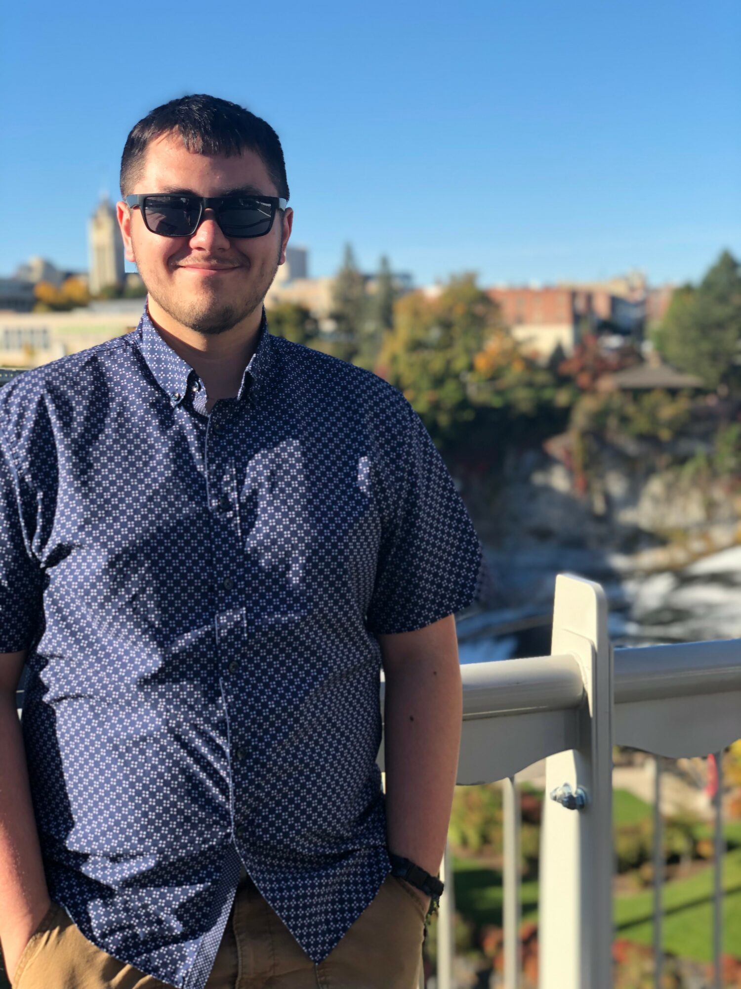 Young man wearing sunglasses and a patterned shirt, standing outdoors with a cityscape and trees in the background.