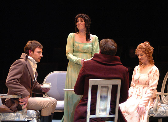 Four people on stage, three seated and one standing, engaged in conversation, with a dark background.