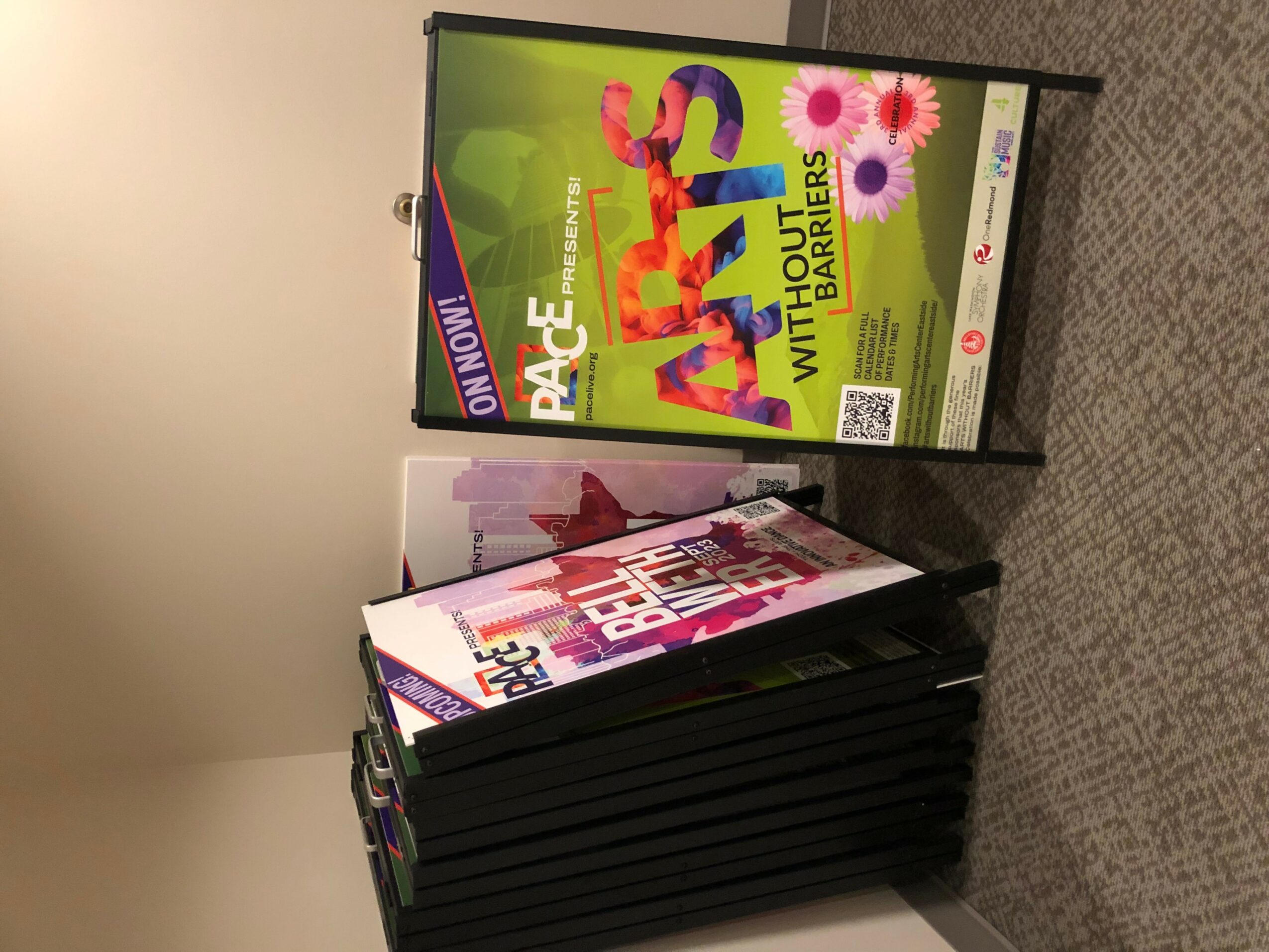 A stack of magazines on a carpeted floor with a digital sign displaying colorful text and graphics behind it.