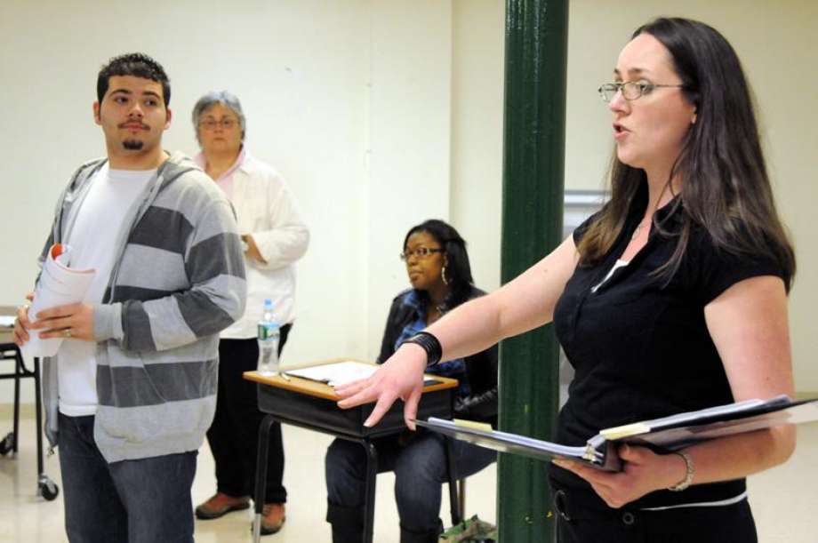 A woman with glasses and dark hair holds a clipboard, speaking to a group of people in a room.