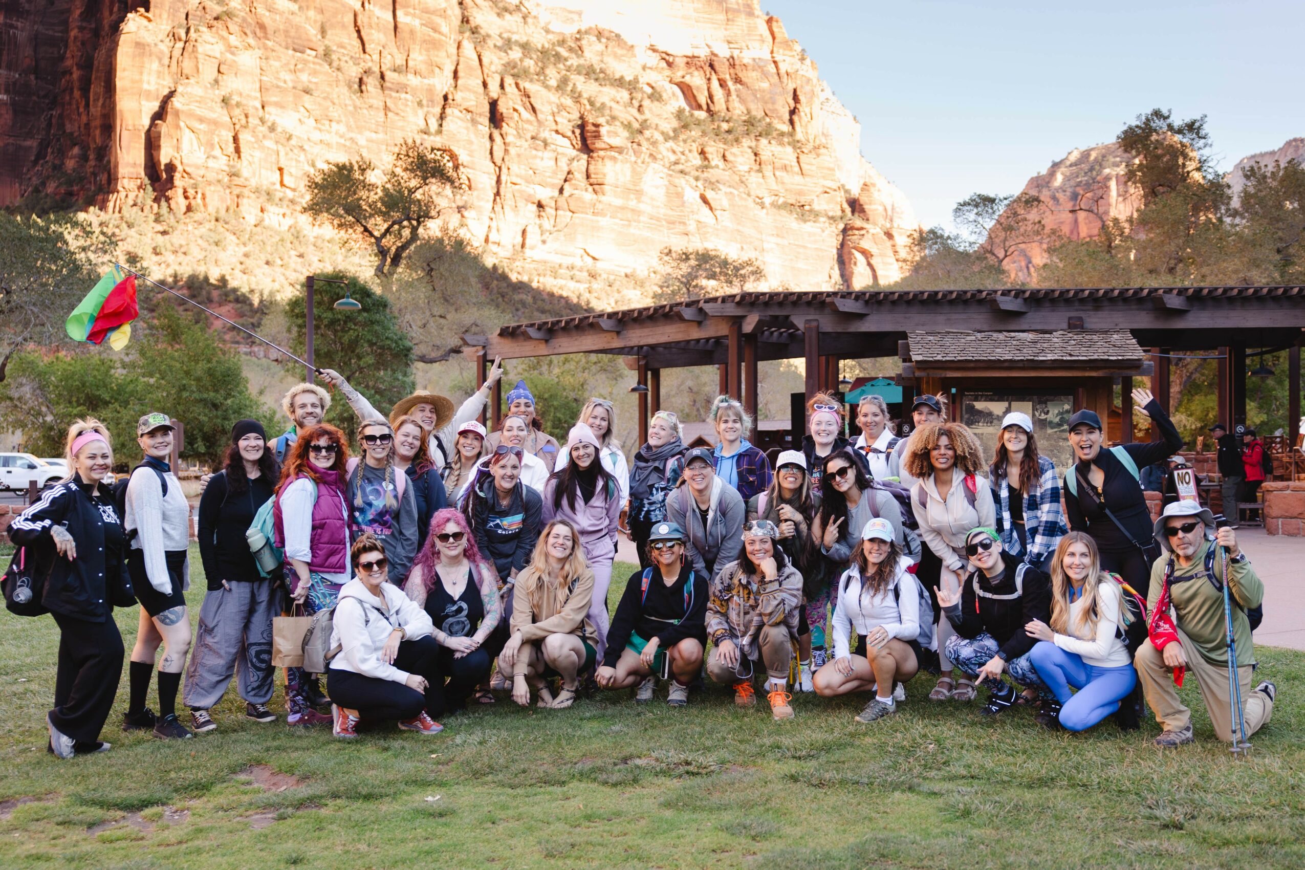 Group of people outdoors in front of a building with rocky cliffs in the background, some holding flags and taking photos.
