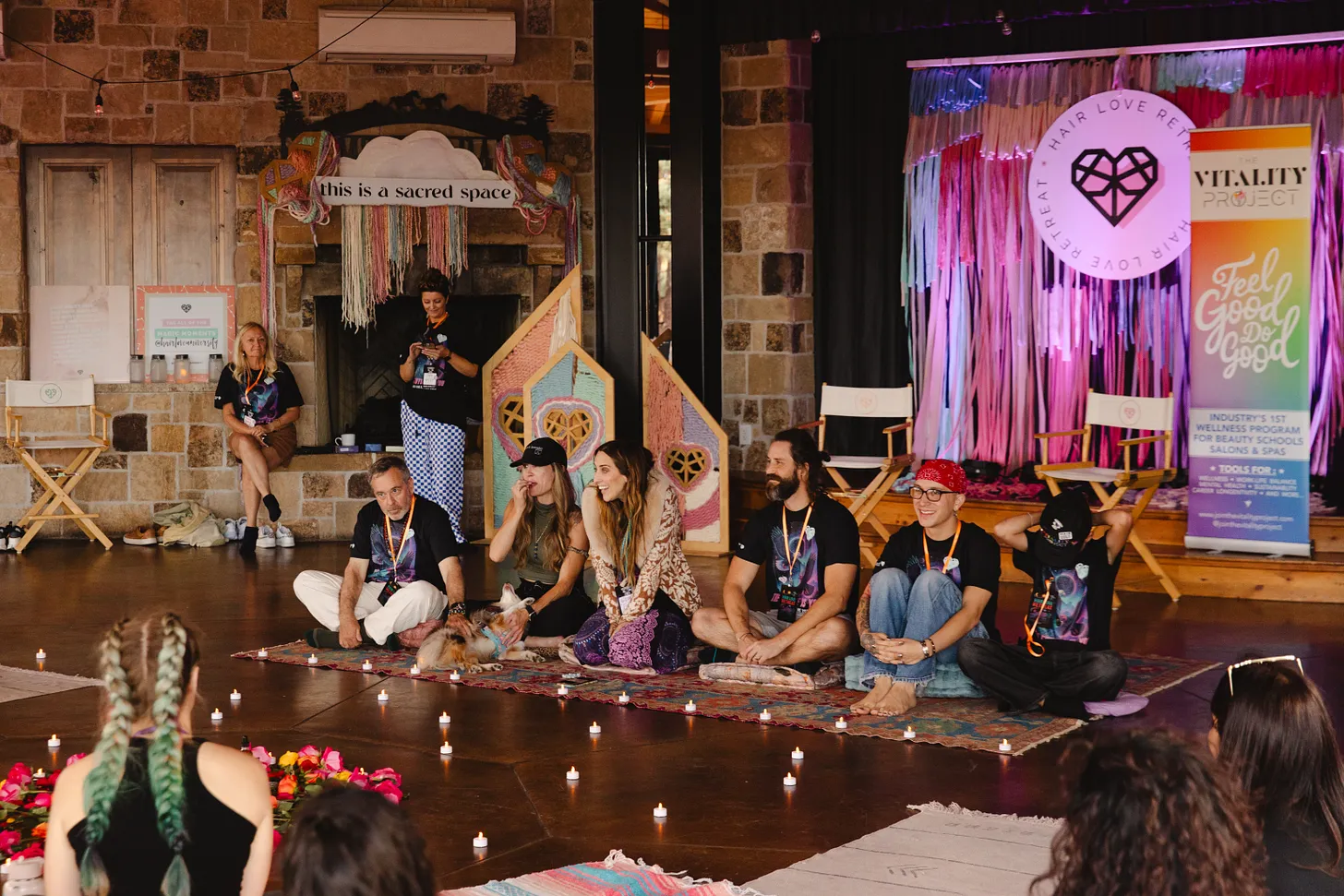 Group of people sitting on the floor in front of a stage with pink curtains and a circular logo, some with microphones.