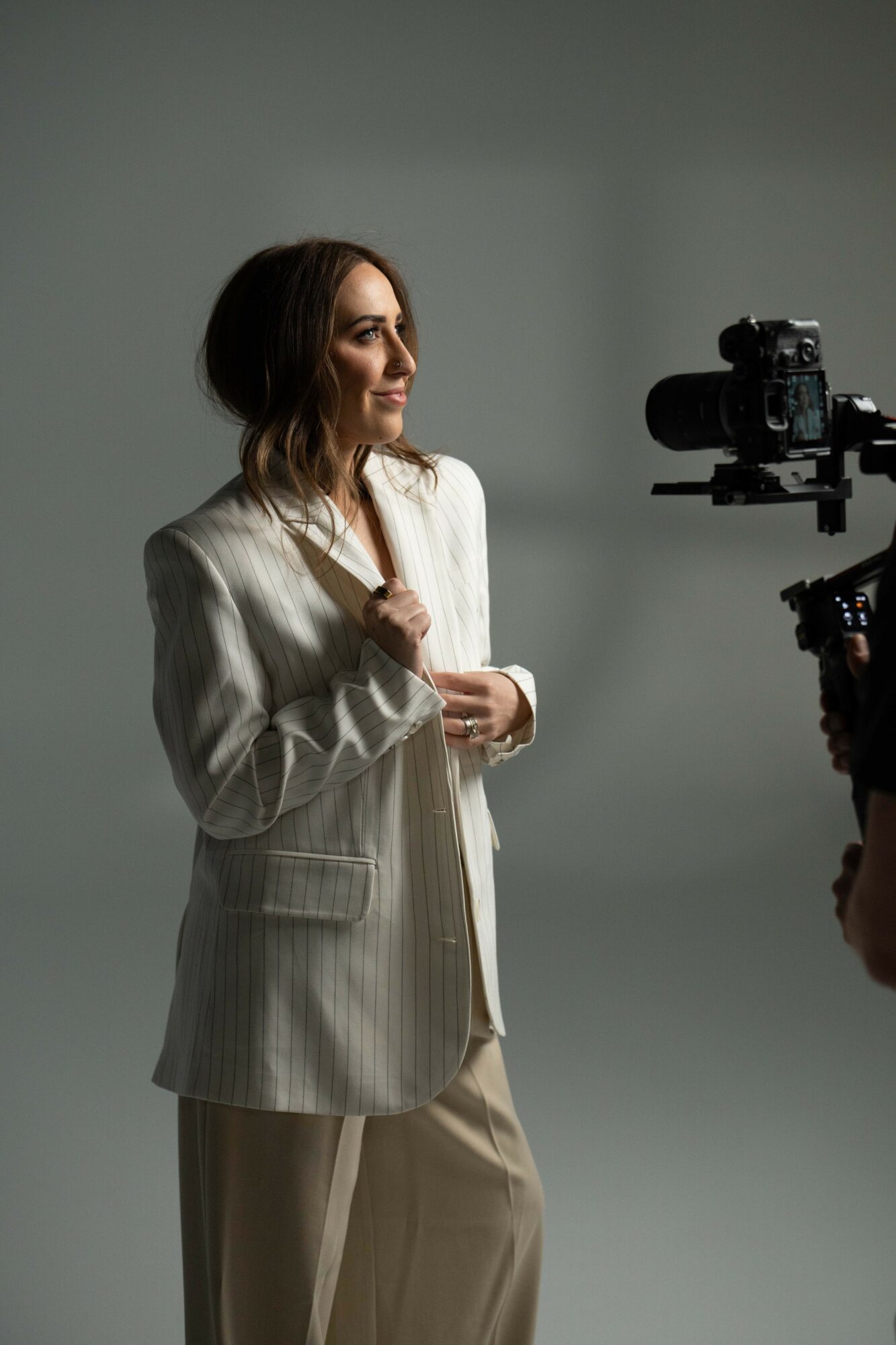 Woman in a light-colored suit being filmed in a studio with a camera, neutral background.