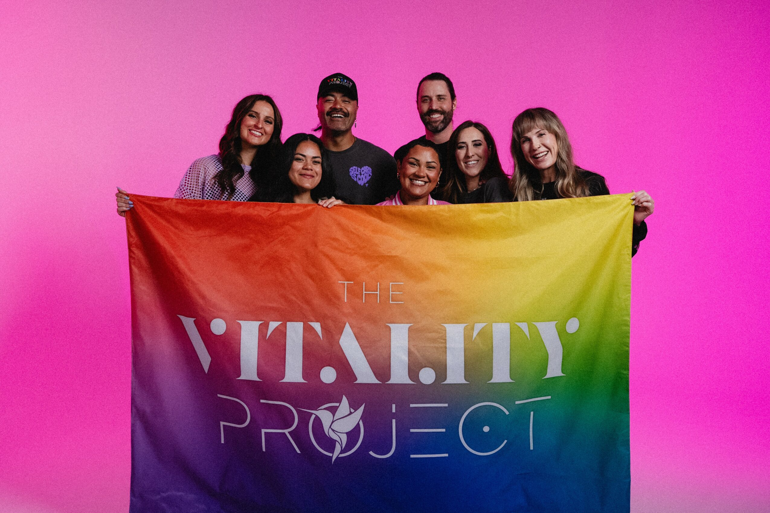 Group of seven people holding a rainbow banner with text, against a pink background.