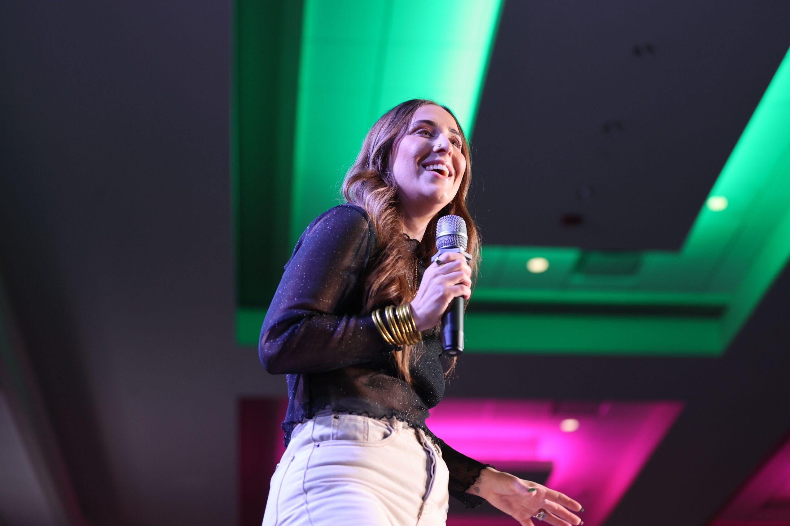 Woman smiling and speaking into a microphone on stage with green and pink lighting overhead.