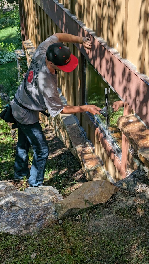 Person wearing a gray hoodie, black cap, and jeans, reaching through a wooden fence with a metal latch outdoors.