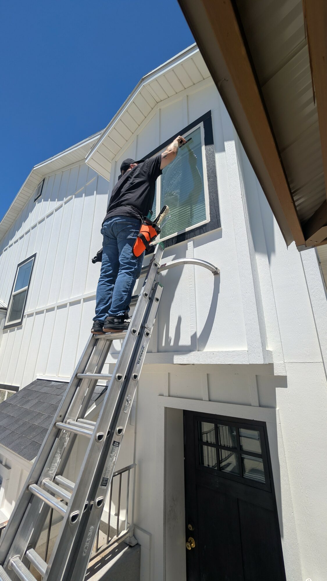 Person standing on ladder cleaning or inspecting a window on a white building exterior, with blue sky background.