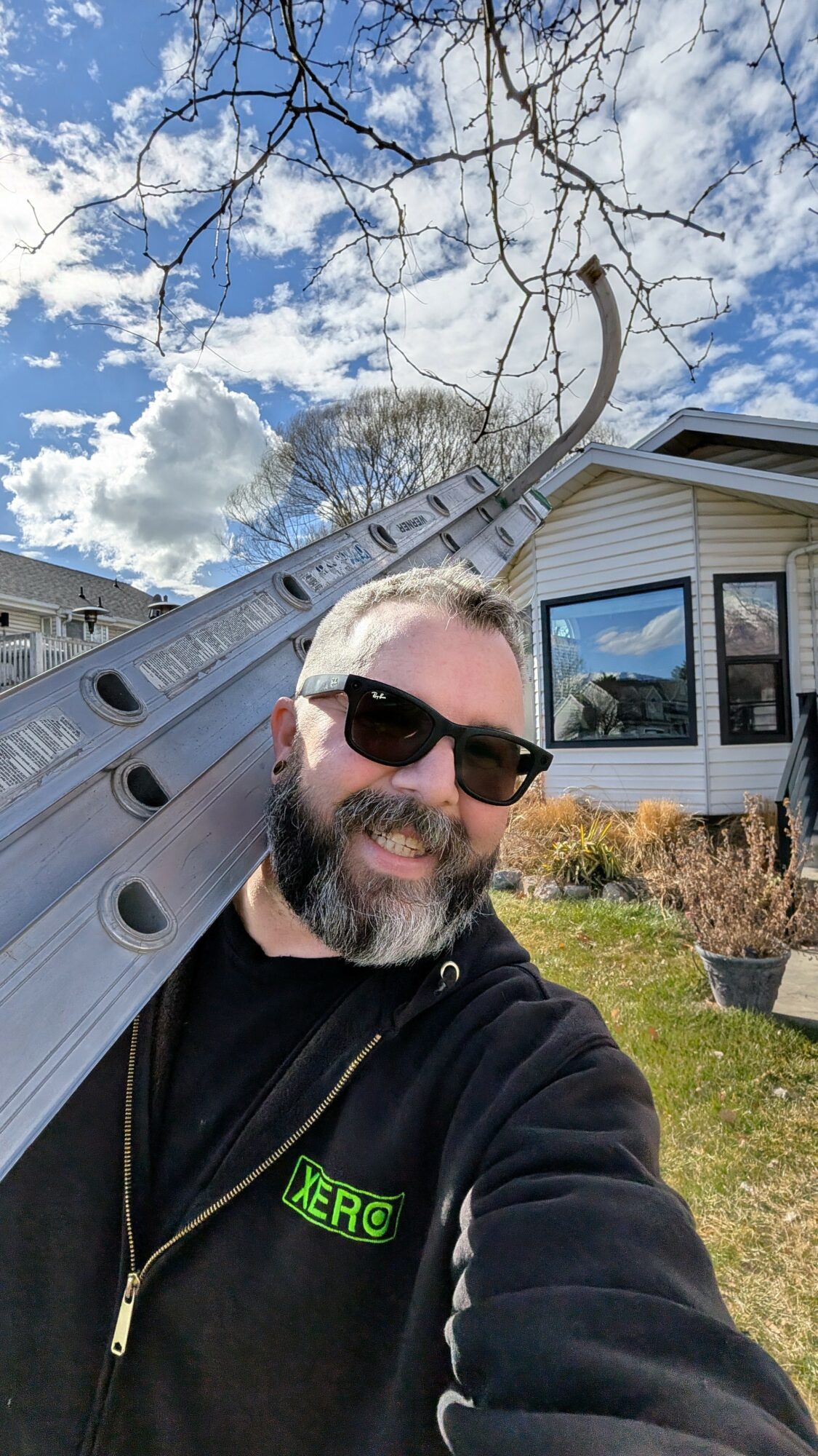 Man with sunglasses smiling outdoors, holding a ladder, in front of a house with a blue sky and clouds.
