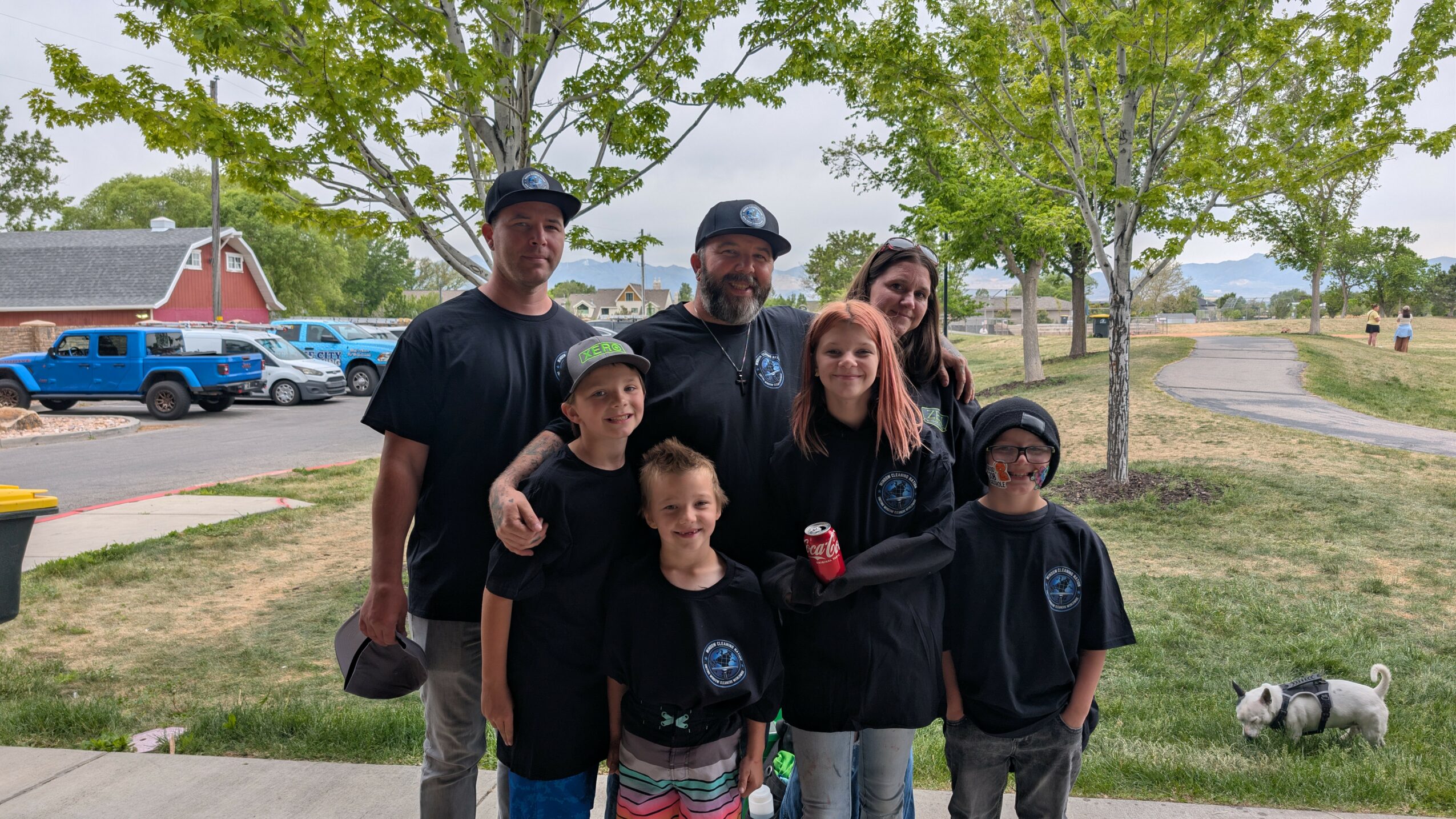 Group of six people, including children, standing outdoors with trees and parked cars in background.