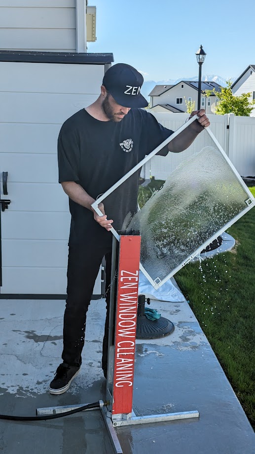 Person cleaning a large glass window outdoors, wearing a black cap and shirt, with a red cleaning tool labeled 'ZEN WINDOW CLEANING'.
