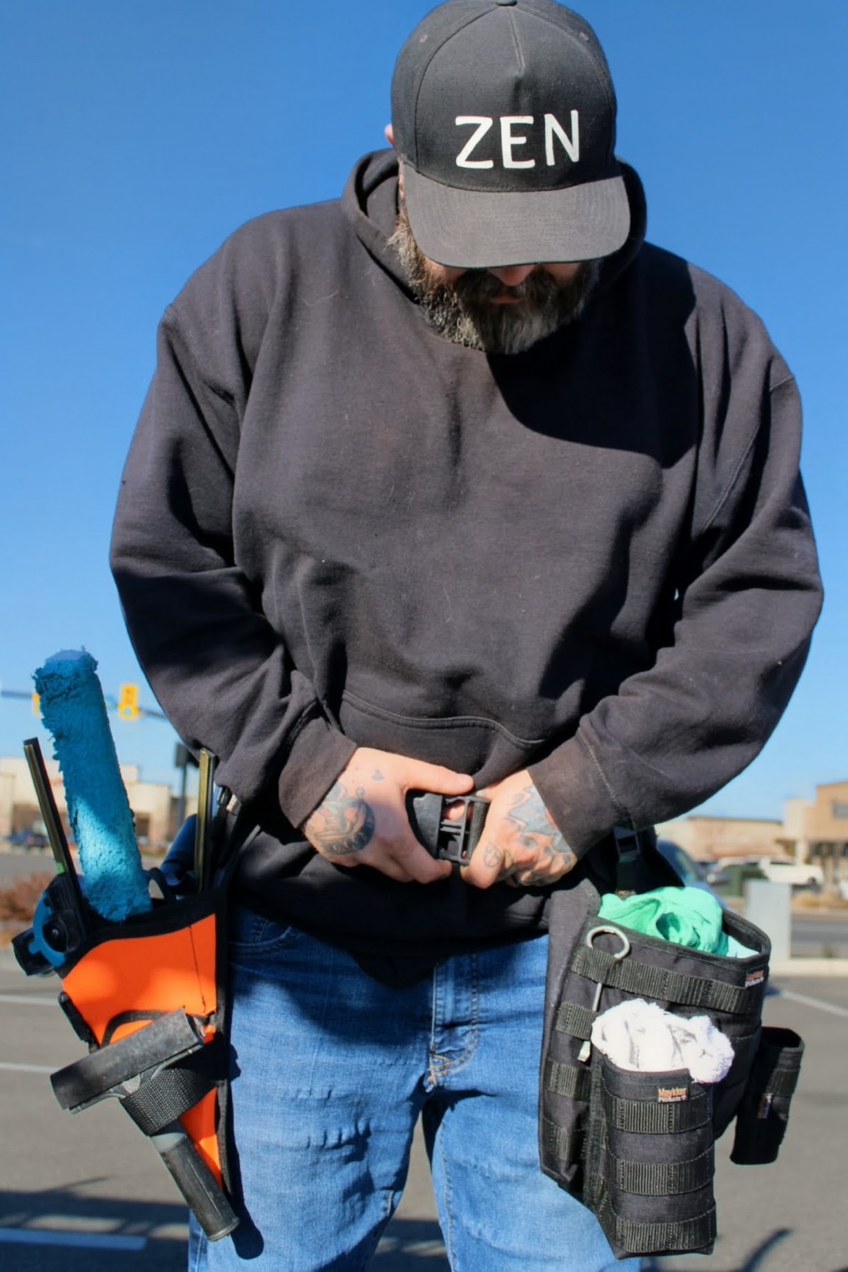 Person wearing a black cap with 'ZEN' text, black sweatshirt, holding a tool, with a tool belt, outdoors against blue sky.