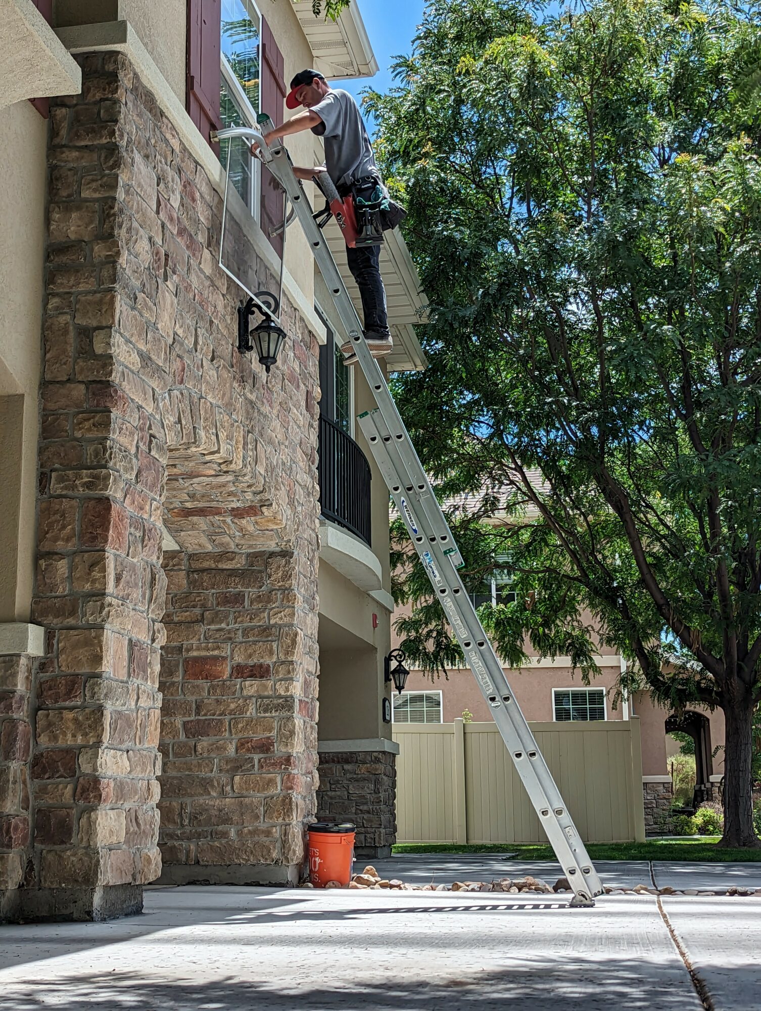 Person on ladder cleaning window outside a house with brick and stone exterior, trees in background, sunny day.