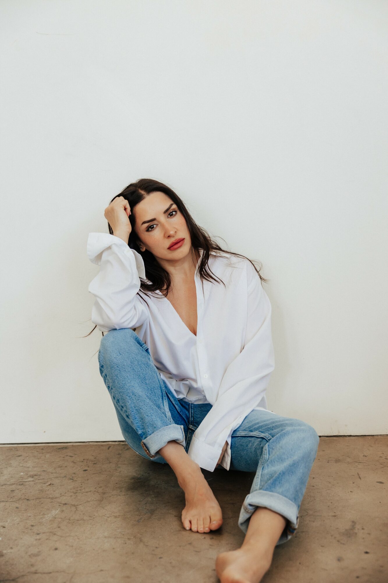 Woman with long dark hair, wearing a white shirt and jeans, sitting on the floor against a plain wall.