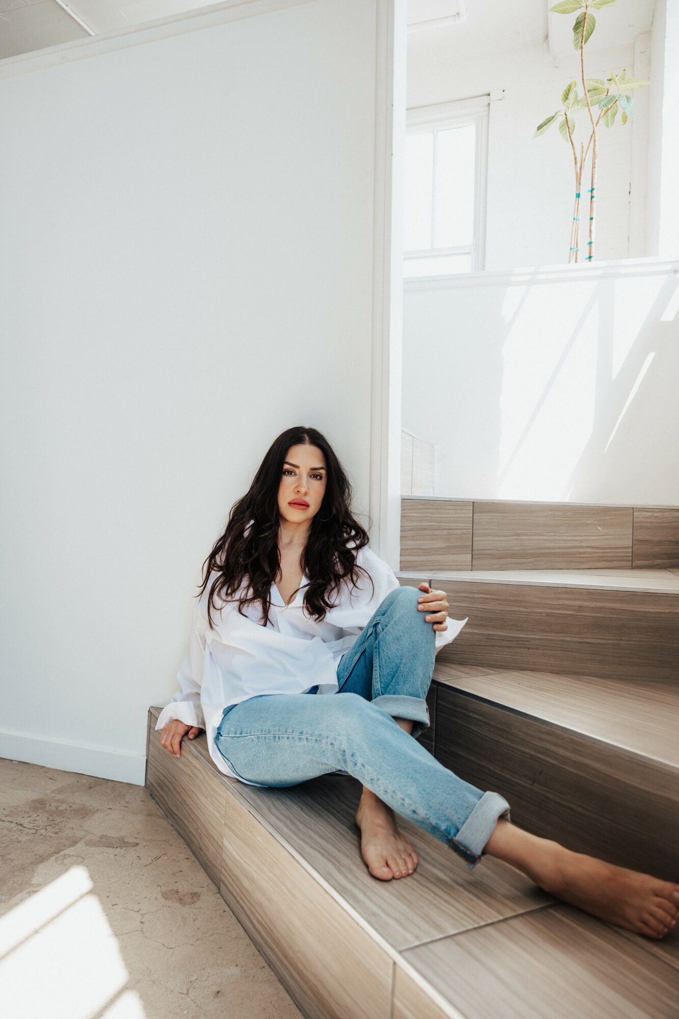 Woman with long dark hair sitting on wooden stairs, wearing a white shirt and jeans, in a bright room with large windows.