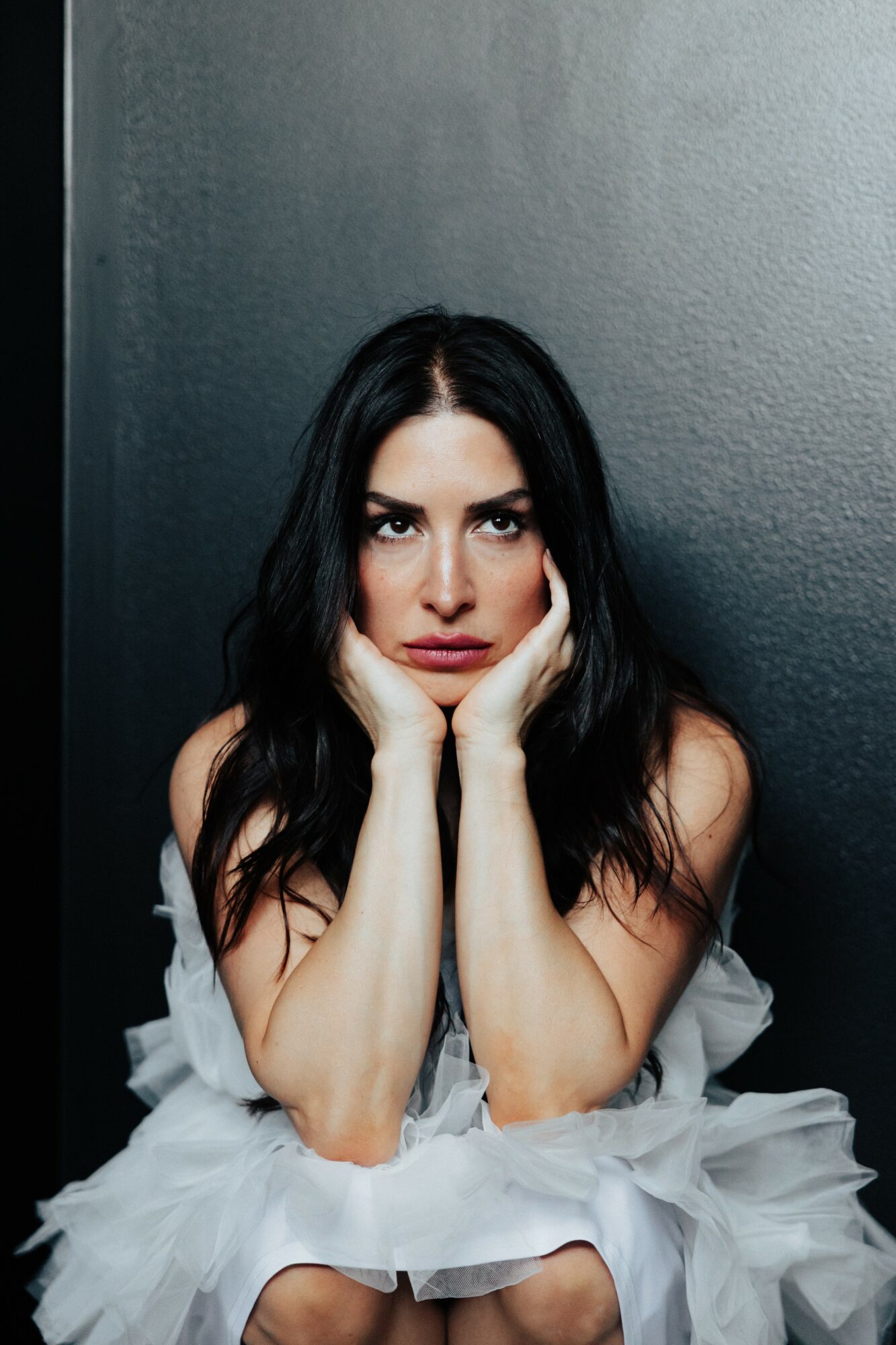 Woman with long dark hair and intense expression, resting her face in her hands, against a dark wall.