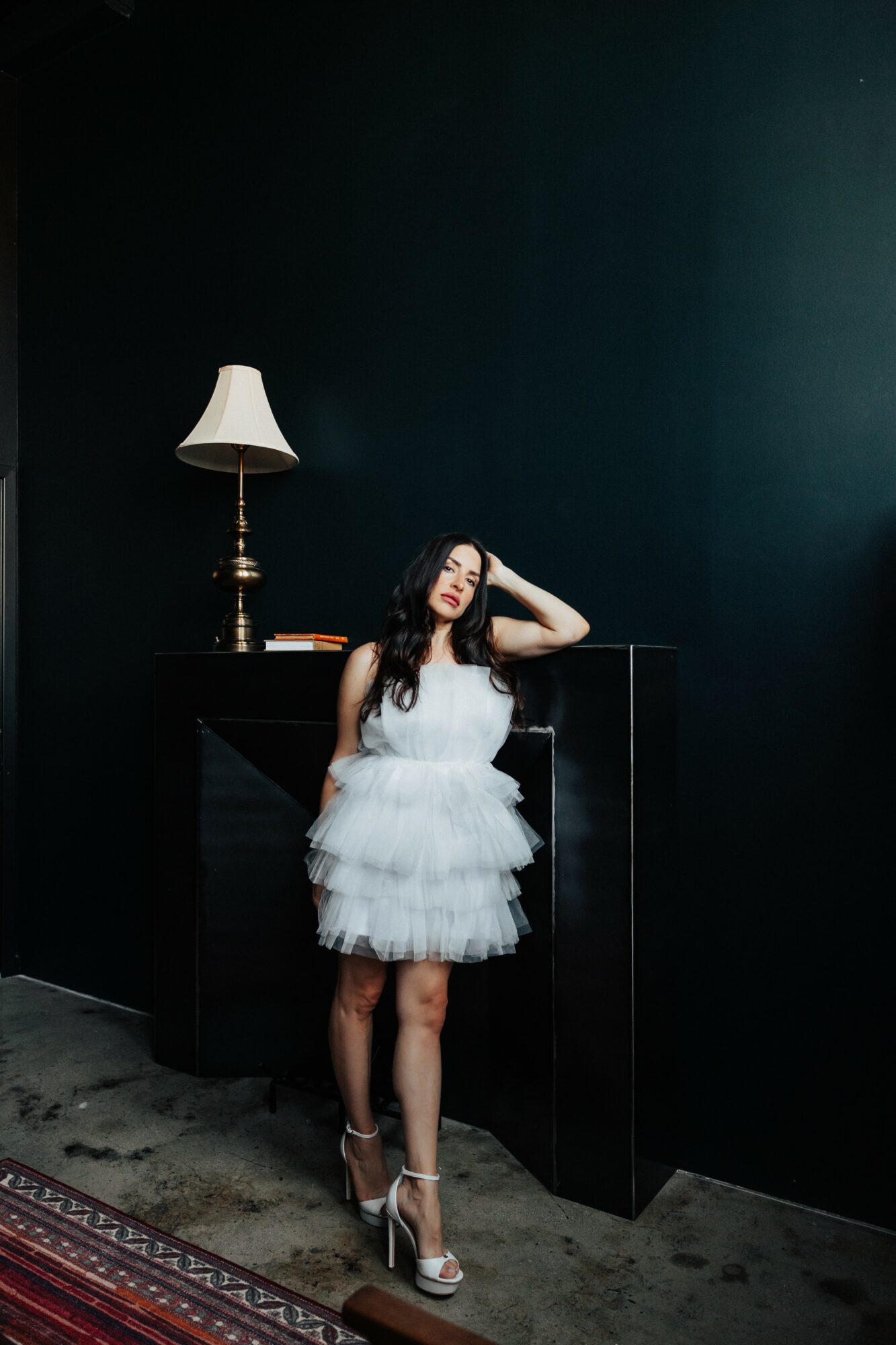 Young woman in a white dress and high heels stands next to a black cabinet with a lamp on top, against a dark wall.