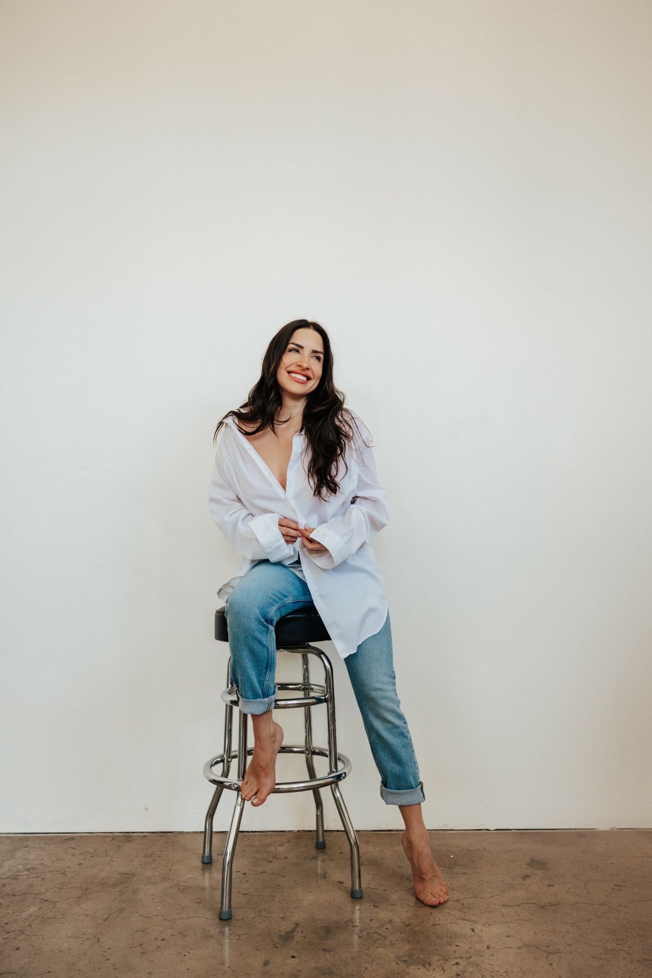 Woman sitting on a stool, smiling, wearing a white shirt and jeans, against a plain background.