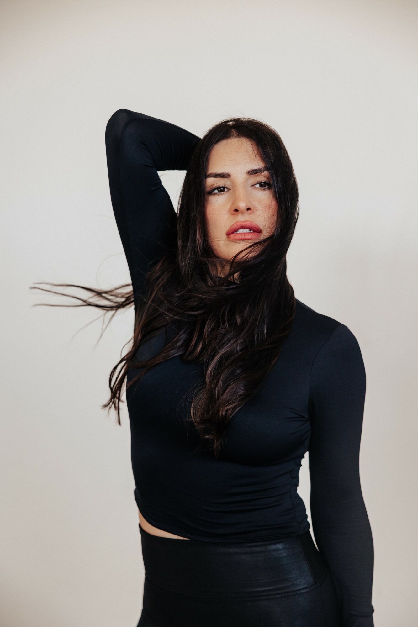 Woman with long dark hair wearing a black long-sleeve top, posing with one arm behind her head, against a plain background.