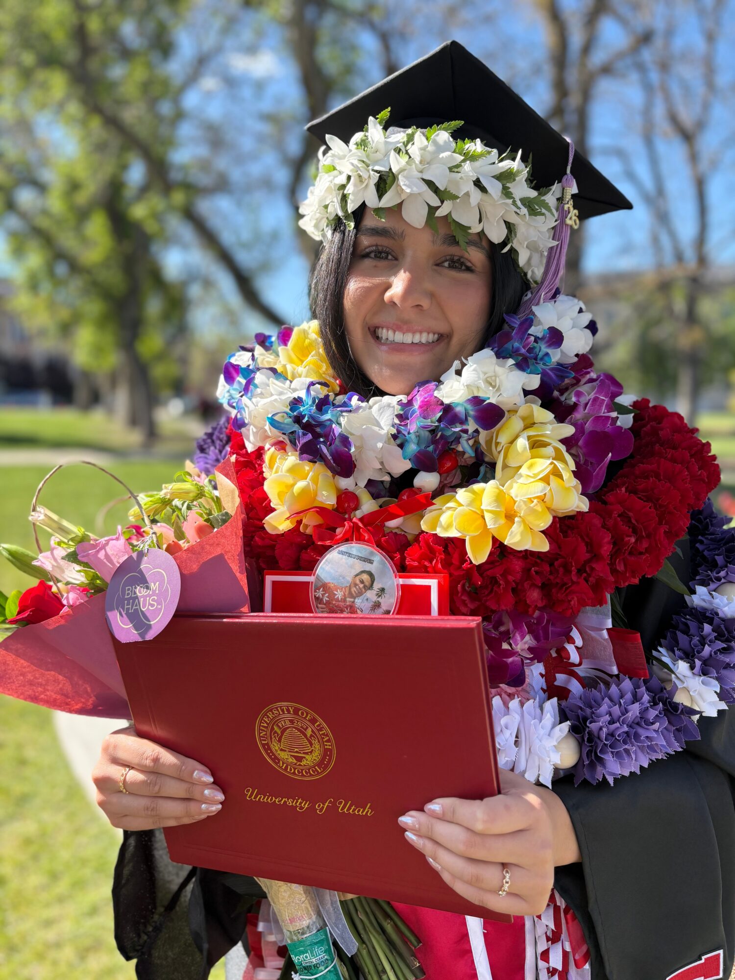 Graduate wearing a cap and flower leis, holding a red diploma folder outdoors.