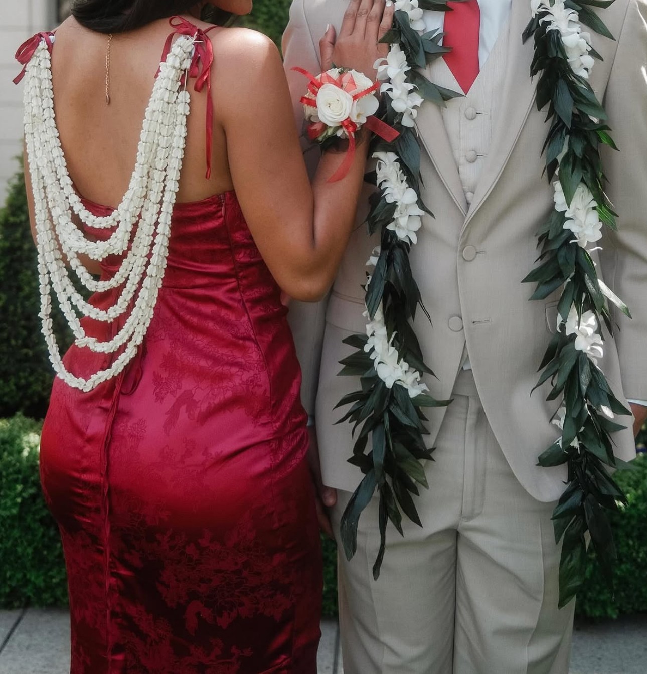 Two people stand together in formal attire with floral garlands; one holds a bouquet.