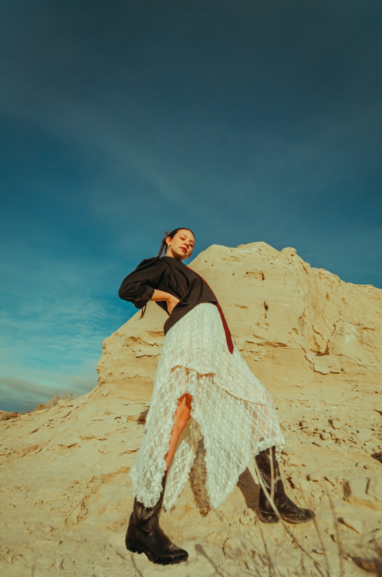 Person standing on sandy terrain with a clear blue sky background, wearing a black top, white skirt, and black boots.