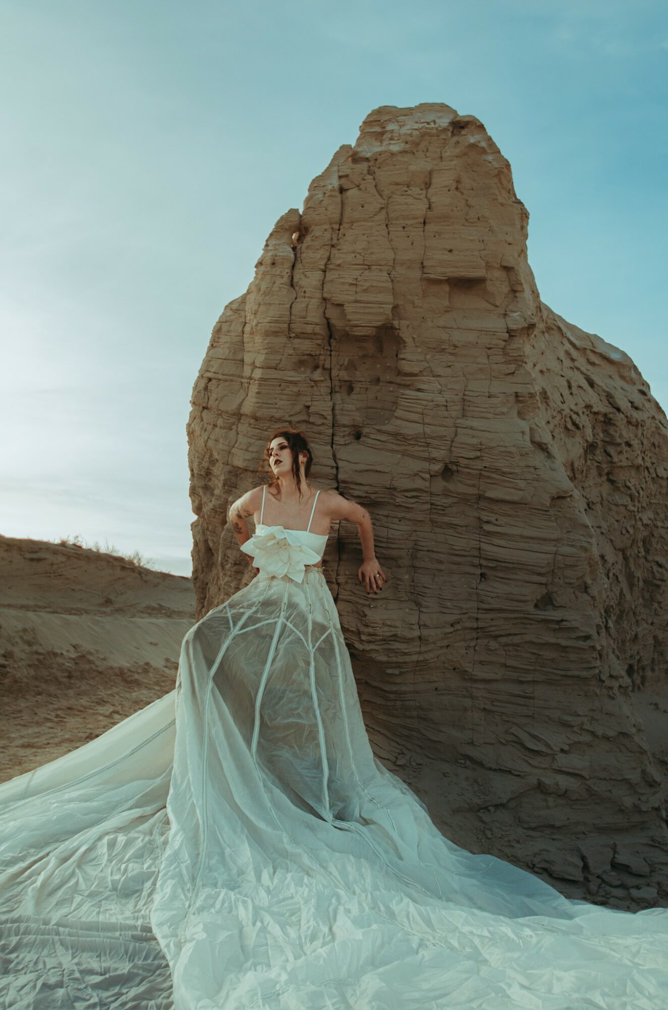 Woman in a white gown stands in front of a large rock formation outdoors, with a clear sky above.