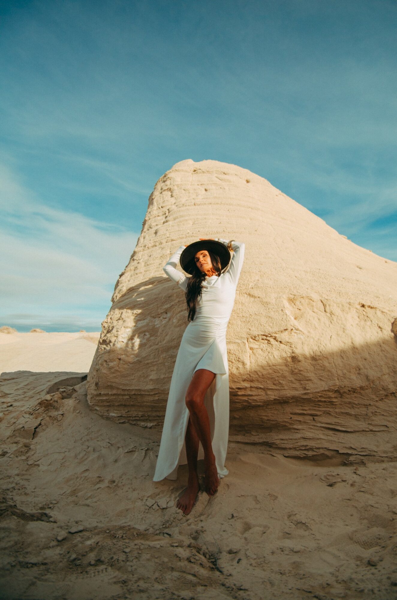 Woman in white dress standing on sandy ground near large white rock formation under blue sky