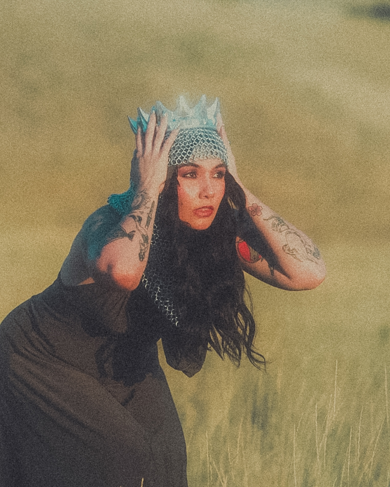 Woman wearing a crown and gloves, standing outdoors with grass background, holding her head with both hands.