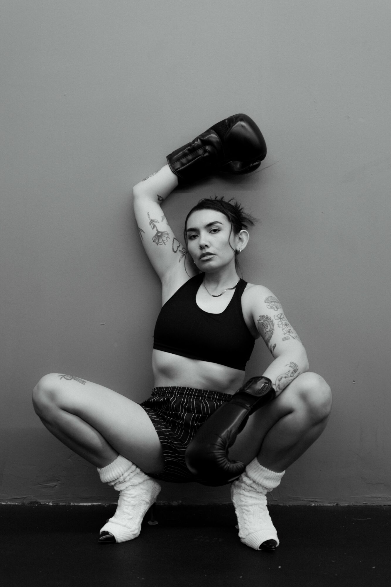 Woman in boxing gloves squats with one arm raised holding a glove, tattoos visible on arms, against plain wall.