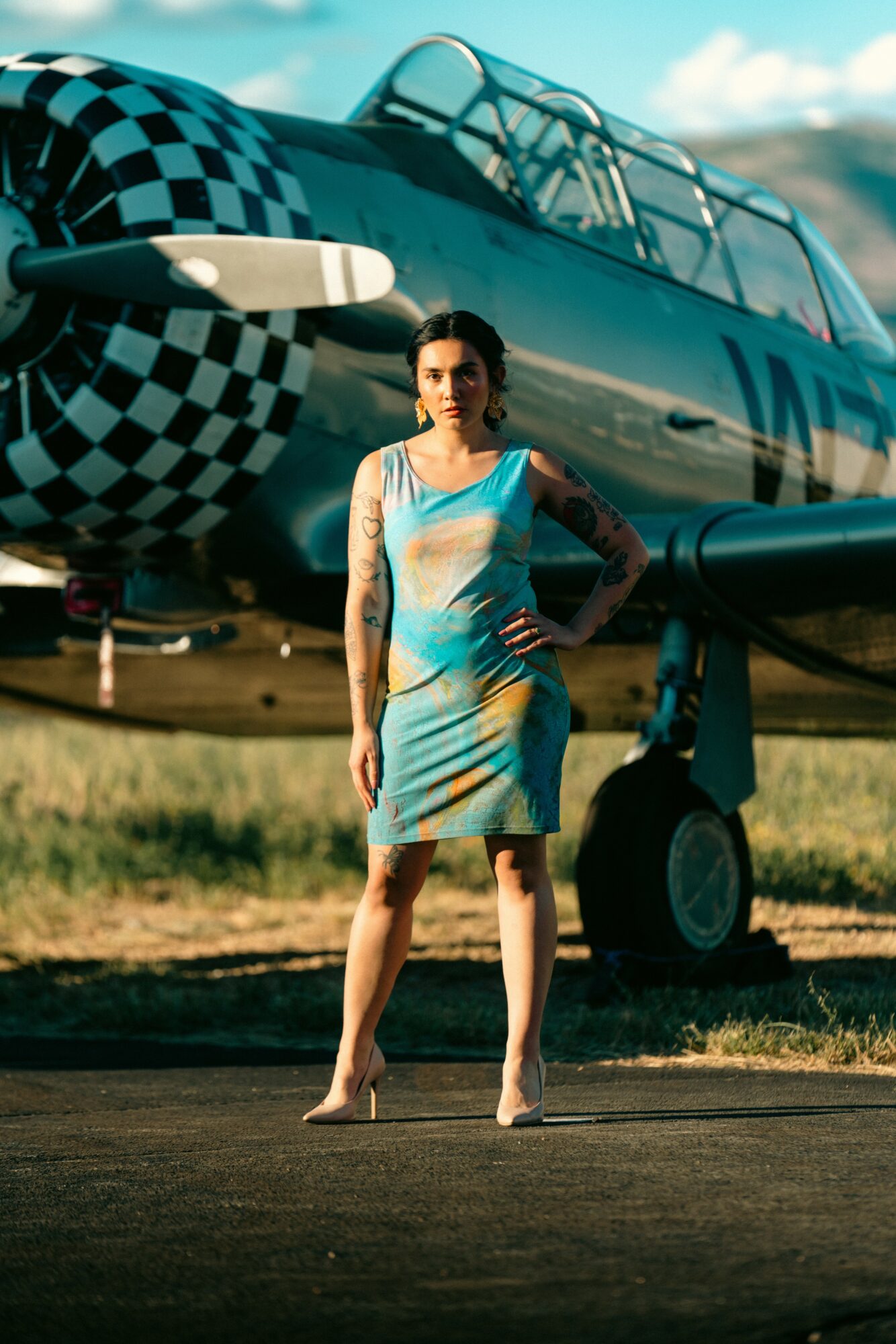 Woman in a colorful dress standing in front of a vintage airplane outdoors, with a grassy field and sky in background.